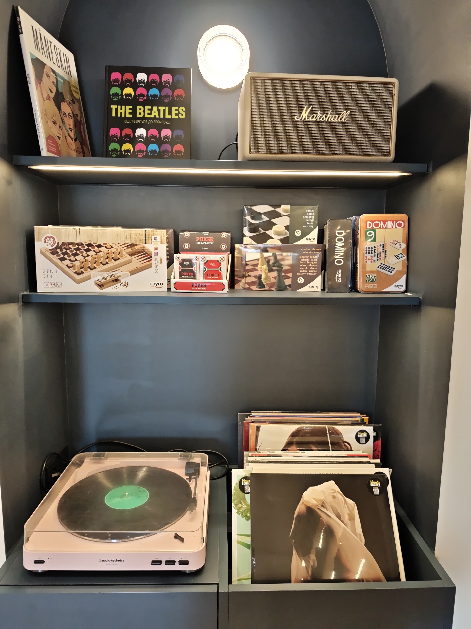 The image depicts a meticulously organized bookshelf in Kyiv, Ukraine, showcasing a curated collection of entertainment and decor items. The top shelf features a "Maneskin" magazine and a black book with colorful Beatles portraits titled "The Beatles: Від Ліверпуля до Еббі-Роуд". Adjacent to these is a beige Marshall speaker, a symbol of audio quality.
The middle shelf is dedicated to board games. A large "Cayro 3 EN 1" set, which includes chess, checkers, and backgammon, is prominently displayed. Beside it are two boxes of "Cayro Poker 100% Plastic" cards and a "Cayro Domino" tin featuring the number 9.
The bottom shelf houses a light pink Audio-Technica record player, with a black vinyl record placed on its turntable. To the right, a stack of vinyl records is neatly arranged, with one album cover prominently displaying a draped figure. The overall environment suggests a contemporary living space, likely a home or a creative studio, emphasizing a blend of modern technology and classic entertainment.
