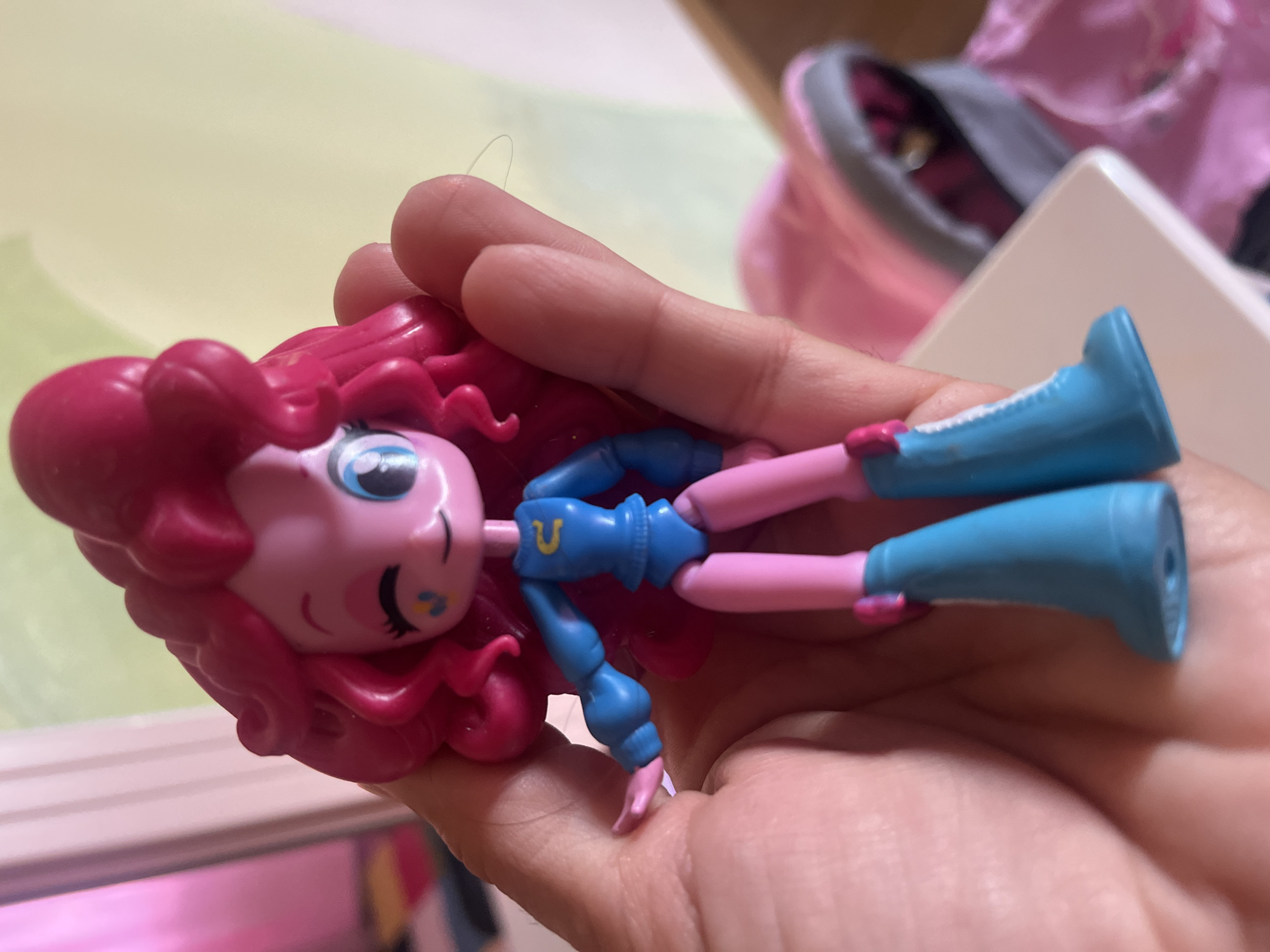 A close-up shot shows a hand holding a pink toy character with bright pink hair and a blue outfit. The toy has a winking eye and a yellow symbol on its chest. It is wearing blue boots. In the background, out of focus, is a pink bag and a wall. The image does not provide any information about the location in the city of Ternivtsi, Ukraine.