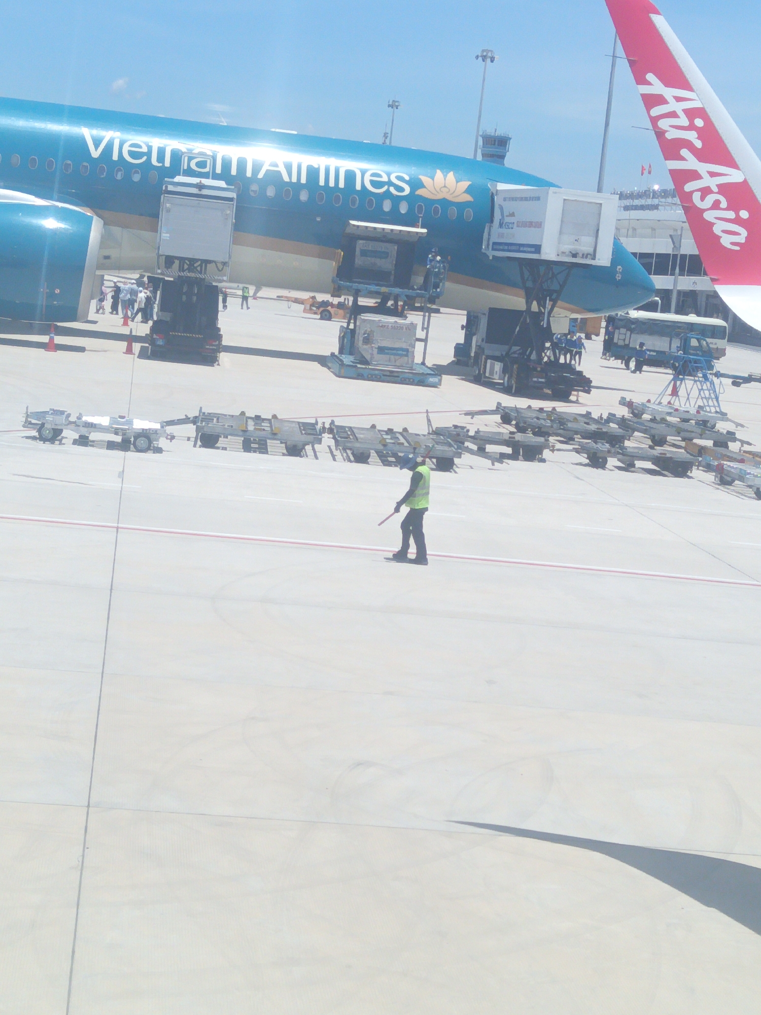 The image depicts an airport tarmac at Cam Ranh City, Vietnam. A large, blue passenger airplane with "Vietnam Airlines" written on its side is docked at a gate. A red and white "Air Asia" logo is visible on the tail of another aircraft in the background. Various airport vehicles, including a baggage loader and a bus, are present around the plane. In the foreground, a person wearing a bright yellow vest and a dark uniform stands on the tarmac, holding a red signaling wand. The ground is paved with concrete, marked with white and red lines. The sky is clear and blue, indicating fair weather. The overall scene captures a moment of aircraft servicing and passenger movement at an airport.