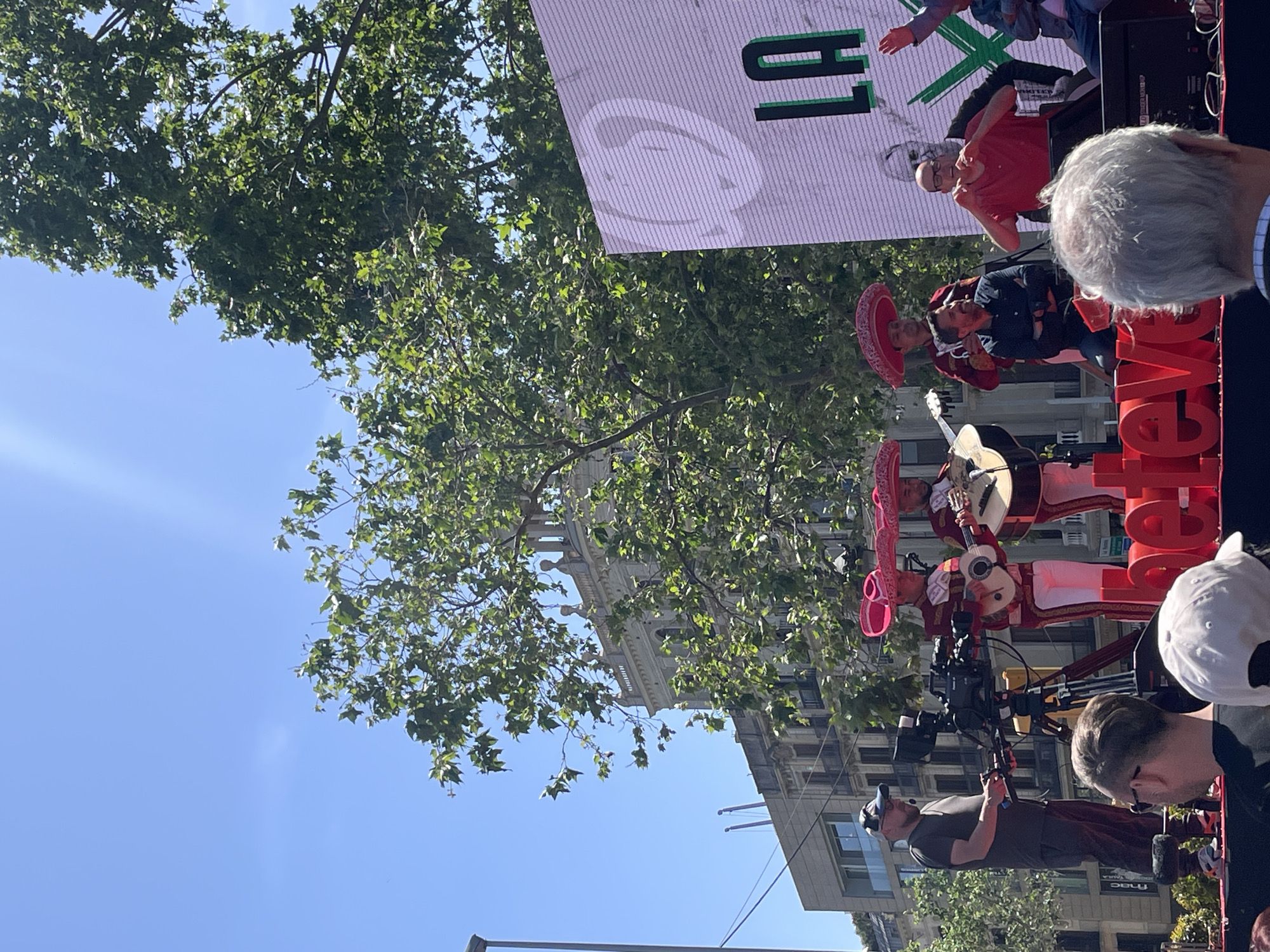An outdoor scene captures a lively event taking place in a city, likely Barcelona, under a bright, clear blue sky during daylight hours. The focal point is a stage or elevated platform where a mariachi band is performing. At least three musicians are visible, dressed in traditional, elaborately embroidered mariachi suits and wearing large pink/red sombreros. Two of them are actively playing instruments, including guitars that appear to be a vihuela and a guitarrón.
To the right of the performers, a large red, three-dimensional sign spells out "teve," possibly part of a longer word like "Esteve." Behind the stage, a large LED screen or banner displays "H1" in green text and a prominent circular logo that resembles a stylized 'C' or 'G'. Also on the stage, a person in a red t-shirt makes a playful gesture with their hands near their face.
In the foreground, several individuals are partially visible. One person with grey hair is seen from behind, as is another wearing a white baseball cap. Below the stage, a camera operator wearing a dark t-shirt and a baseball cap is focused on the event, operating a professional video camera mounted on a tripod.
The background features lush green trees, suggesting a park or a tree-lined street. Beyond the foliage, various city buildings are visible, including older structures with classical architectural details and more modern facades, with some text like "FNA" (potentially "FNAC") faintly visible on one building. The presence of mariachi music, camera crew, and a large screen suggests a public cultural event, festival, or broadcast.
