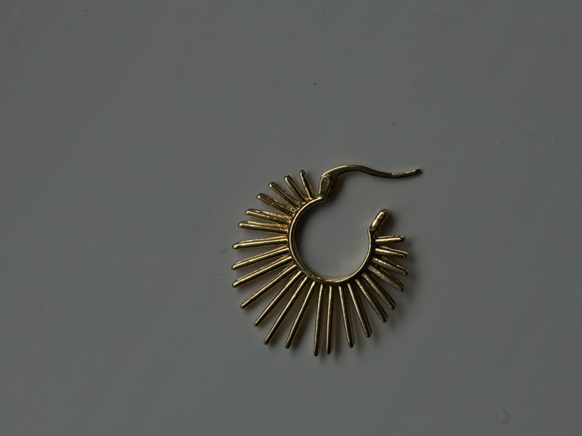 An overhead shot captures a singular gold-colored ear cuff on a flat, gray surface. The ear cuff is designed to resemble a sunburst, with numerous pointed rays radiating outwards from a curved central piece that is open at the bottom. The surface appears to be a smooth, solid material, possibly a table or a floor. The lighting creates soft shadows around the ear cuff, highlighting its metallic sheen and intricate details. The background is a uniform gray with no discernible features or markings. There are no people or other objects visible in the frame. The image is cropped to focus primarily on the ear cuff, with a significant amount of negative space surrounding it. There is no information available to determine the location in the city of Chirchik, Uzbekistan.