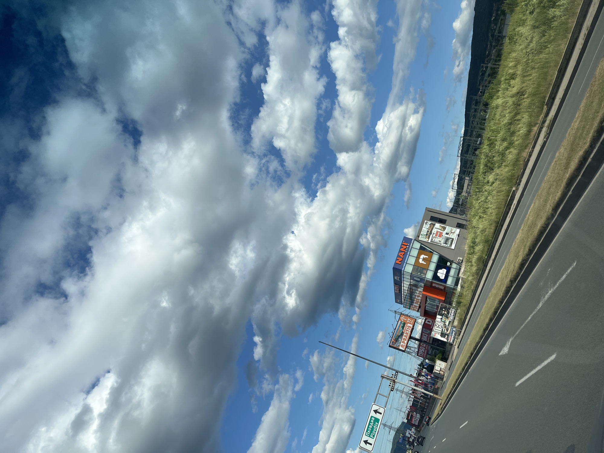 The image captures a view from a moving vehicle on a highway, looking towards a commercial area under a cloudy sky. In the foreground, a two-lane road with white markings curves to the right. A grassy median separates the road from the adjacent land. To the left, a green road sign indicates "Plovdiv" with an arrow pointing upwards. Beyond the median, a cluster of buildings is visible, including multi-story structures with prominent signage for "NANI HOME" and "PRAKTIS 17." Several flags are also displayed in front of the buildings. In the background, rolling hills and power lines are discernible. The sky is filled with a mix of bright blue patches and large, white and gray clouds.