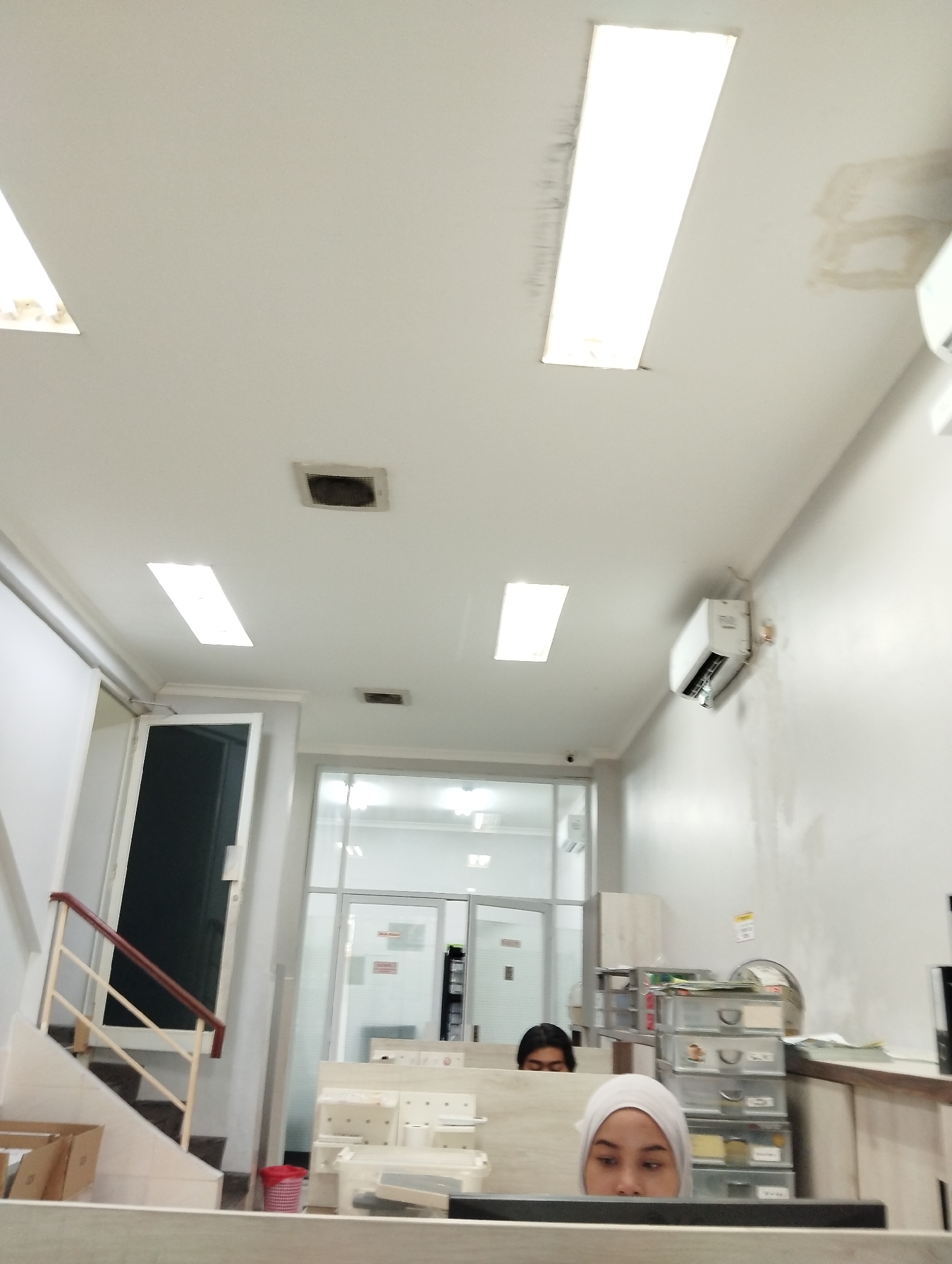 The image depicts an indoor scene, possibly an office or administrative area, in Kelapa Dua, Indonesia. The ceiling features recessed rectangular lights and some water stains. An air conditioning unit is mounted high on a white wall. In the foreground, a woman wearing a white hijab is looking down. To her left, a man with dark hair is partially visible behind a partition. Behind them, there are white shelves with various items, including clear plastic drawers filled with what appear to be supplies or documents. To the left of this area, a staircase with a red and beige railing leads down. A cardboard box is visible at the bottom of the stairs, and a red and white striped trash can is nearby. The background shows glass-paneled doors and windows, suggesting additional rooms or an entrance.