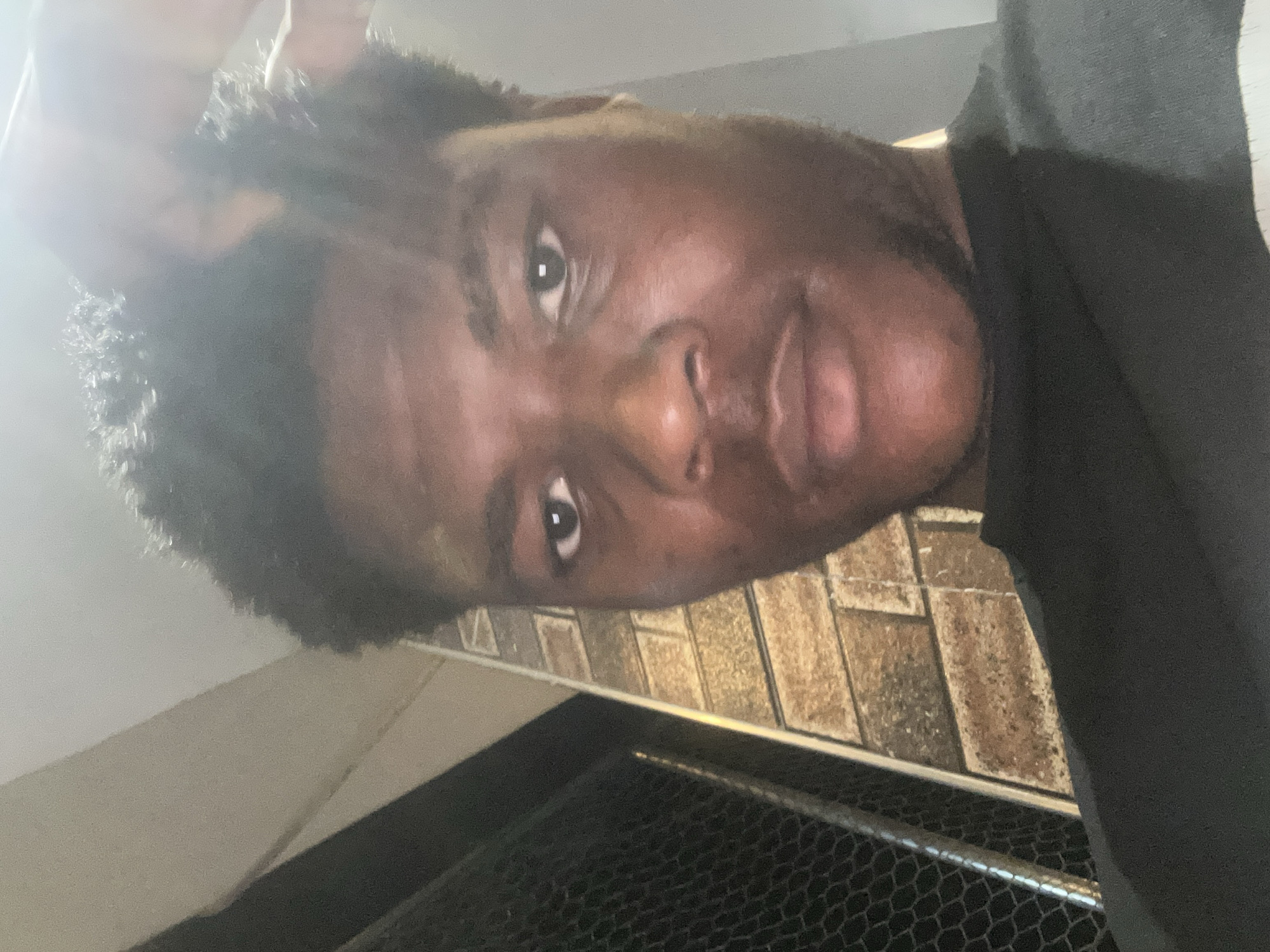 A close-up, waist-up shot shows a Black male, likely in his late teens or early twenties, with dark, curly hair and dark skin. He is wearing a black t-shirt and is positioned slightly to the right of the frame, looking directly at the camera. His right hand is raised and touching his hair. The background consists of a brick wall with a textured, light brown and gray pattern, and a dark, lattice-like fence to the left. The lighting suggests an outdoor evening setting, possibly in Ihiagwa, Nigeria, as indicated by the provided context.