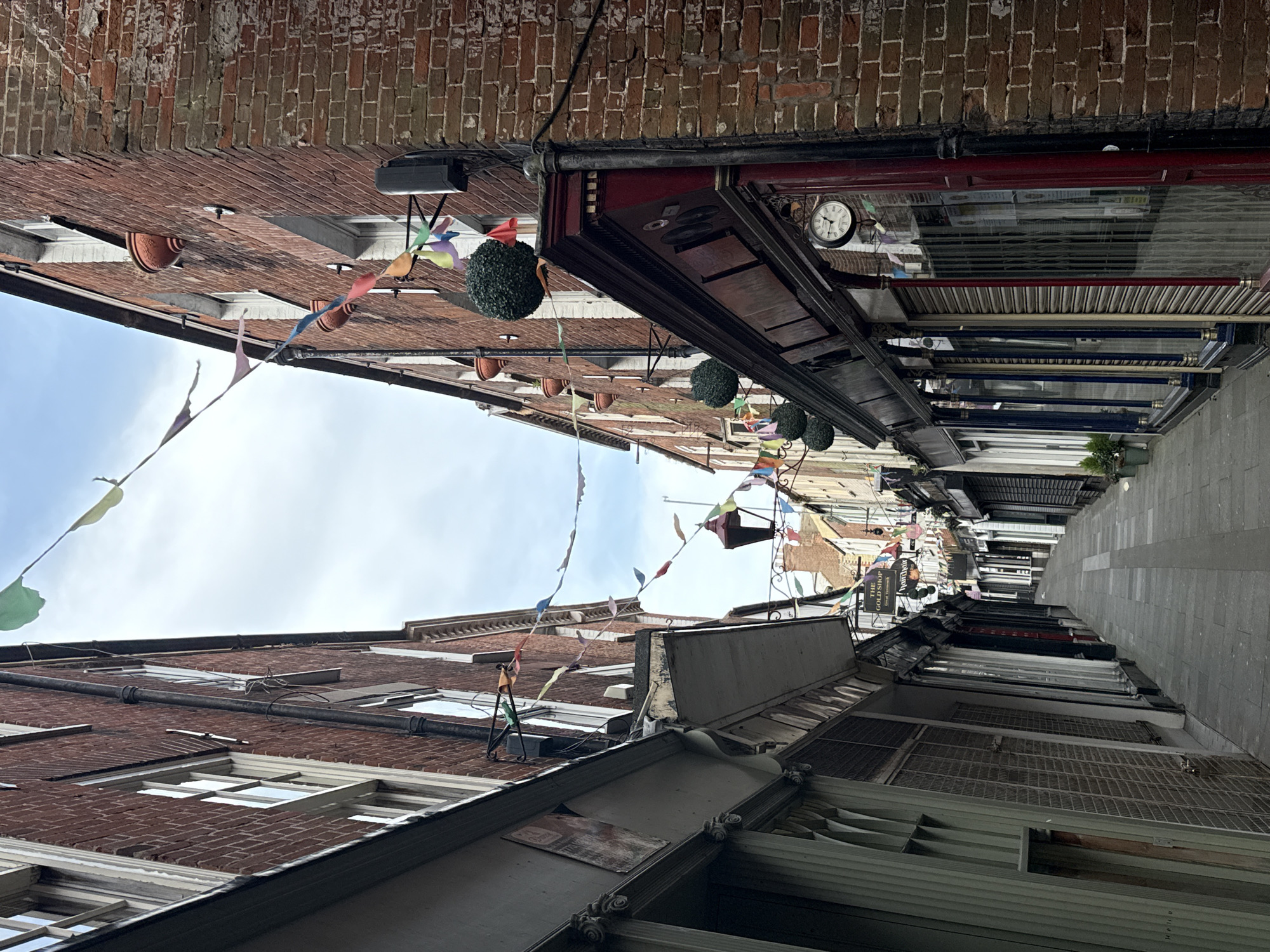 The image depicts a narrow pedestrian alleyway in Great Yarmouth, United Kingdom. The view is oriented upwards, emphasizing the tall brick buildings lining both sides of the street. Colorful pennants are strung across the alley, adding a festive atmosphere.
On the right side of the alley, shopfronts are visible, some with closed metal security shutters. One shop has a clock mounted above its entrance. A narrow grey-paved walkway runs through the alley.
The left side shows similar brick buildings with some visible windows. The sky above is partially cloudy. The alley appears mostly deserted.