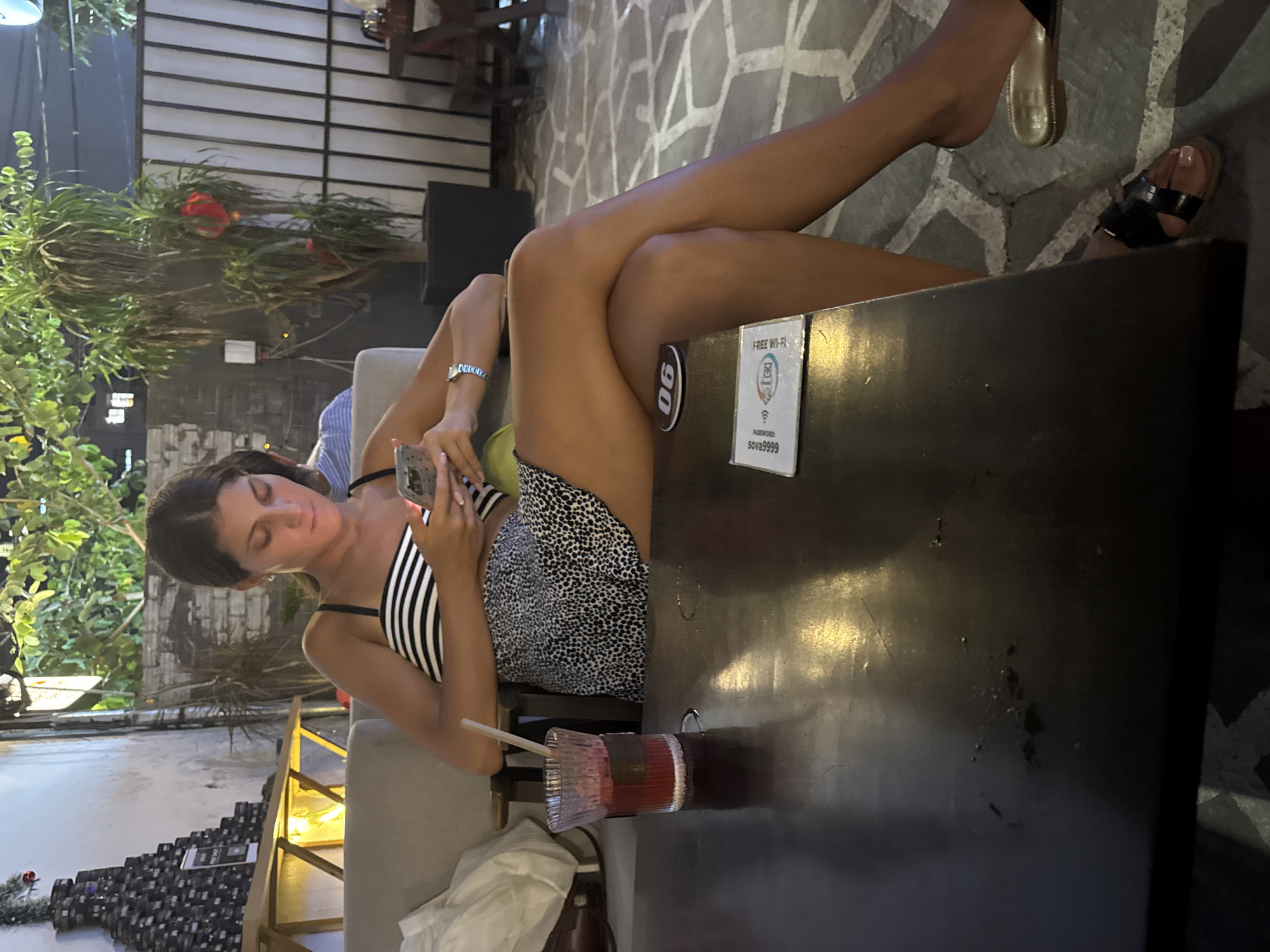 The image depicts a woman sitting at a table in what appears to be an outdoor café or restaurant. She is fair-skinned with dark hair, wearing a striped and dotted pattern dress with thin straps. She is looking at her phone in her hand, suggesting she might be texting or browsing. A cocktail with a straw is on the table next to her. The table is dark, and a sign indicating "Free Wi-Fi" is attached. Her legs are crossed over the table, with one foot wearing a slide and the other wearing black sandals on the stone ground. The environment suggests a warm, possibly tropical location, given the greenery and outdoor setting. While the location is not evident from the photo, it has been mentioned as being Đà Nẵng, Vietnam.