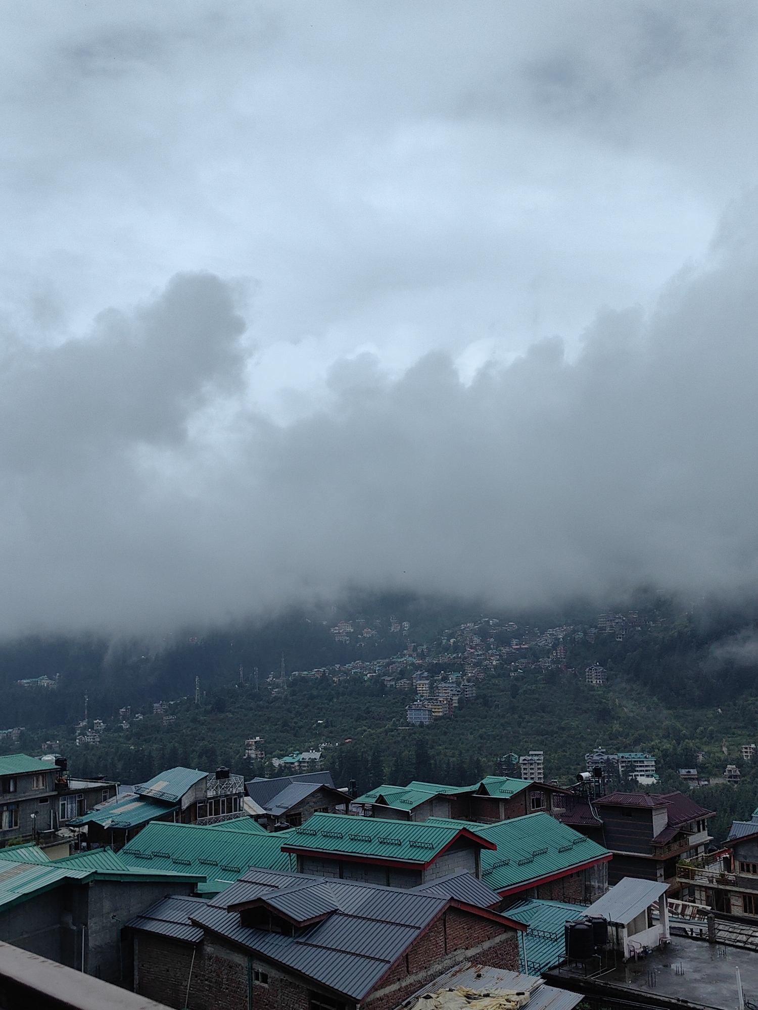 This image depicts a hazy, overcast view of the town of Vashisht, India, nestled in a valley. In the foreground, a cluster of buildings with distinctively green and gray corrugated metal roofs are visible. These structures appear to be residential or commercial, with varying architectural styles. Some buildings are multi-storied, while others are smaller, single-family dwellings.
The midground and background show the town sprawling up a lush, tree-covered hillside. The density of buildings increases as they ascend the slopes. The upper portion of the image is dominated by heavy, grey clouds that obscure the sky and add to the atmospheric haze. The overall scene conveys a sense of a mountainous region experiencing cloudy or misty weather, likely during the monsoon season given the lush greenery. No people are visible in the image. The image is a still photograph, implying a static scene with no ongoing actions or interactions.