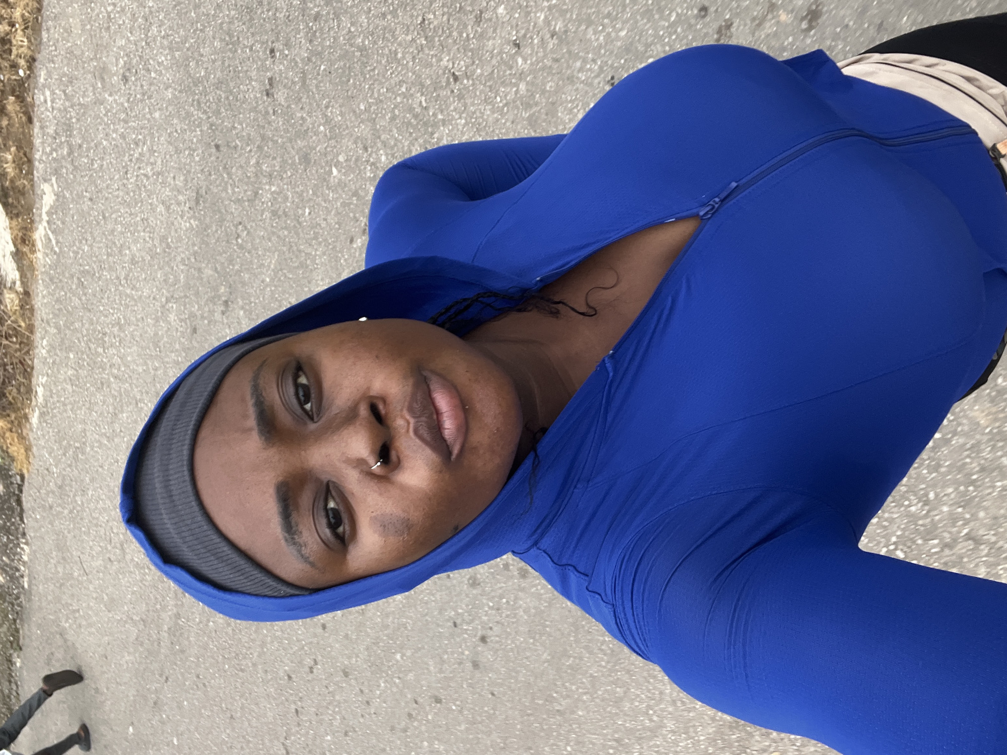 The image shows a medium shot of a woman from the chest up, facing the camera and looking slightly to her right. She is wearing a vibrant royal blue hooded jacket with a zipper that is partially open. Underneath the jacket, a grey beanie is visible on her head. She has dark skin, a nose ring, and some skin discoloration on her left cheek. Her hair is styled in dark braids that are partially visible.
The background is an asphalt surface, likely a road or pathway, with some dry vegetation and gravel visible along the edge. In the upper left corner of the image, a person's legs and feet are visible, walking away from the camera, wearing dark trousers and brown shoes.
The image appears to be a selfie taken outdoors, possibly during a walk or exercise session. While the location is stated to be Abuja, Nigeria, there are no specific landmarks or indicators within the image to confirm this definitively. The overall lighting suggests an overcast day or the shade from an overhead structure.