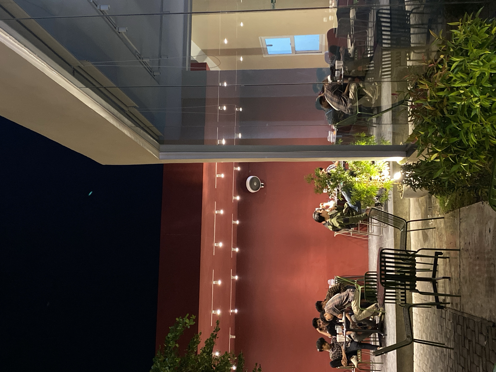 The image depicts an evening scene at a cafe or restaurant with both outdoor and semi-indoor seating areas, likely in Lhokseumawe, Indonesia. The dominant feature of the outdoor section is a vibrant deep red wall illuminated by multiple rows of horizontal string lights, casting a warm glow. Several individuals, appearing to be young adults, are casually seated at dark metal tables and chairs in this outdoor space. They are seen socializing, with some engaged in conversation or looking at their phones. Green potted plants are strategically placed, adding natural accents to the concrete patio floor.
A large glass partition divides the outdoor area from an interior space, which is primarily visible through reflections on the glass. These reflections show more people seated, a light-colored ceiling with recessed lighting, and a distinct rectangular window or skylight emitting a blue light. The reflections create a layered visual effect, blending the exterior and interior views.
The time of day is clearly night, indicated by the dark sky visible on the left side of the frame and the reliance on artificial lighting from the string lights and interior fixtures. A white circular object, possibly a speaker or an exhaust vent, is mounted high on the red wall. The scene conveys a relaxed and social atmosphere. No discernible text is clearly visible in the image.