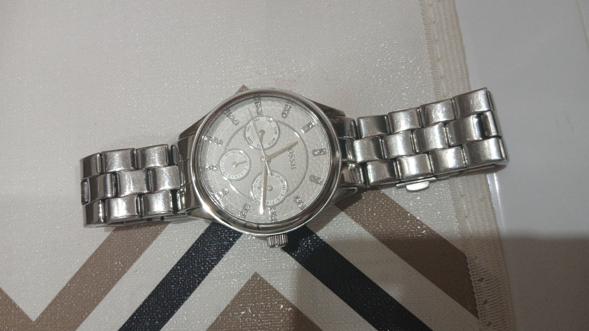 A silver-toned wristwatch with a metal band is shown in a top-down view, lying on a textured surface with diagonal stripes of brown, black, and white. The watch face is circular with a textured silver background and features three subdials. The central dial displays "TISSOT" written horizontally. The subdials show information related to days of the week, dates, and possibly month or year. Small, diamond-like markers indicate the hours. The watch band is made of interlocking rectangular links. The background surface appears to be a placemat or tablecloth.
