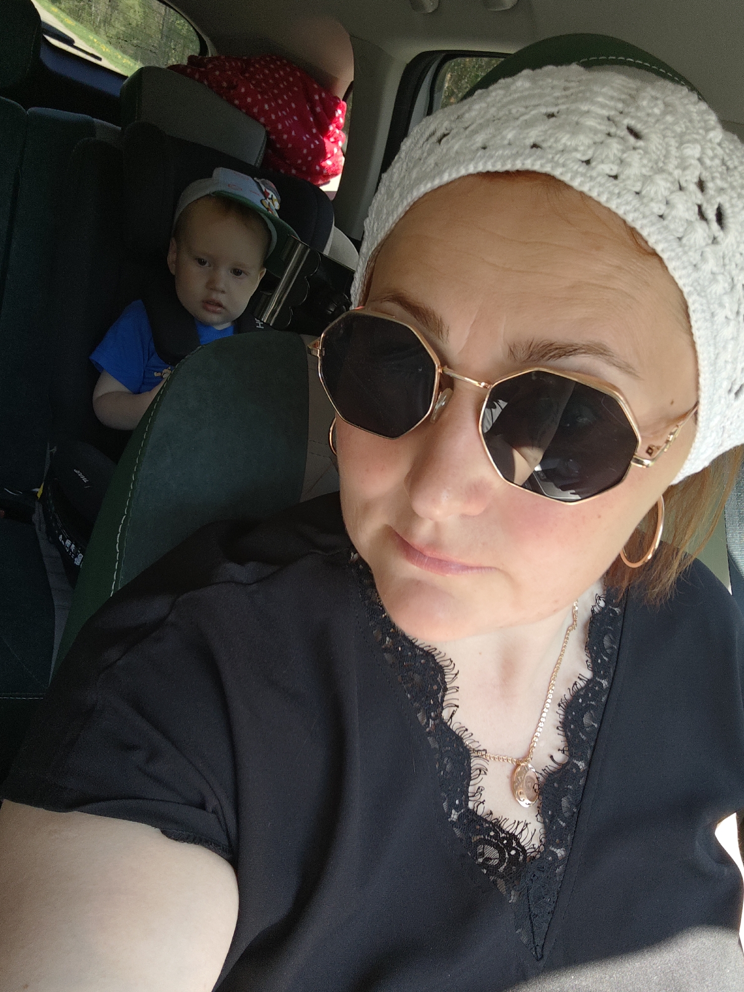 The image shows a woman in the foreground wearing a white crocheted hat, sunglasses, and a black shirt with lace trim, along with gold hoop earrings and a gold necklace. Behind her, in what appears to be the back seat of a car, a young child is seated in a car seat, wearing a blue shirt and a baseball cap. Another person with a red and white polka dot shirt is also partially visible in the background. The scene appears to be inside a vehicle, likely in transit. Without further context, I am unable to determine if the location is Chaikovsky, Russia.