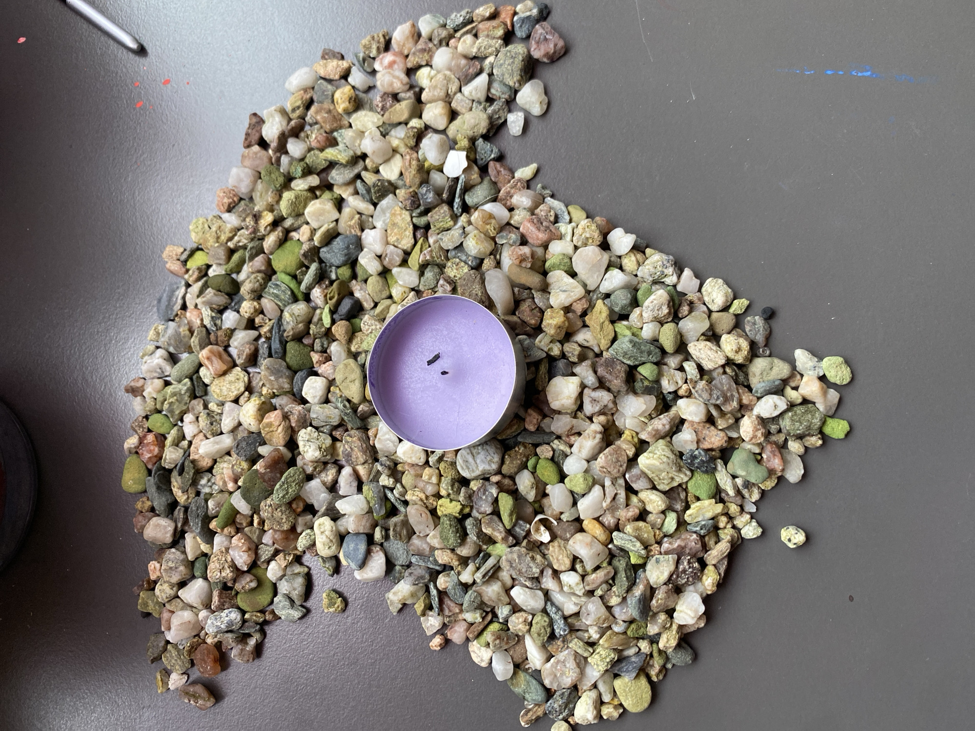 The image shows a pile of colorful pebbles arranged in a somewhat heart-like shape on a dark gray, textured surface. A small, lavender-colored tea light candle sits in the center of the pebble arrangement. The pebbles are of various sizes and colors, including shades of green, gray, white, brown, and pink. Some of the pebbles are smooth, while others are rough and irregular. The candle is cylindrical and made of wax, with two small black wicks visible. The surface the pebbles and candle are on appears to be a tabletop or a similar flat surface. In the upper left corner of the image, the tip of a silver pen or stylus is visible, along with a few small red smudges. Along the top edge of the gray surface, there are faint blue markings, suggesting it might be a surface used for art or crafts. The lighting suggests it is daytime, with diffused light casting soft shadows. The overall scene is static and appears to be a still life arrangement. There is no indication of people, weather, or specific time of day beyond general daylight. The location context of Bílina, Czechia, does not provide any visual cues within the image itself. No discernible text is present in the image.