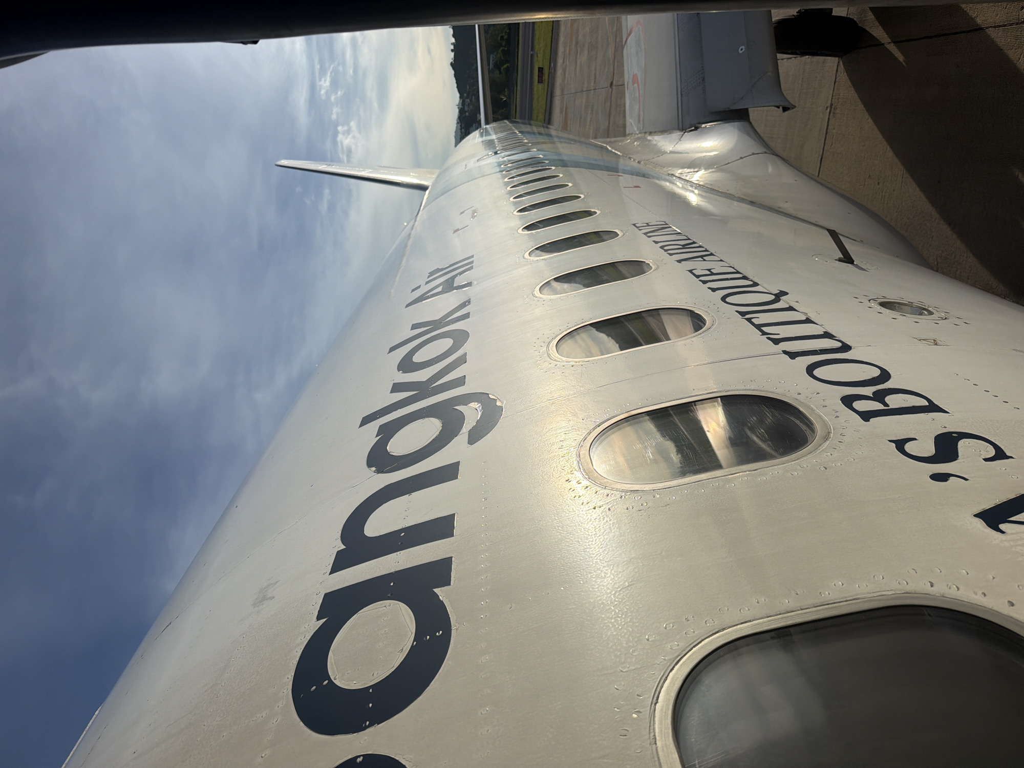 This is a close-up, high-angle shot of the fuselage of a narrow-body aircraft, likely on the ground at an airport. The aircraft is primarily white with dark blue lettering. The text visible on the fuselage reads "Bangkok Air" and "Boutique Airline". Several oval windows line the side of the plane, reflecting the sky and clouds. The sky appears partly cloudy, suggesting it is daytime. Some dew or moisture is visible on the surface of the aircraft. The image is taken from a perspective that suggests the viewer is either inside the aircraft looking out or standing very close to it on the tarmac. The overall impression is of an aircraft preparing for or having just completed a flight, with the text indicating a connection to Bangkok, Thailand.