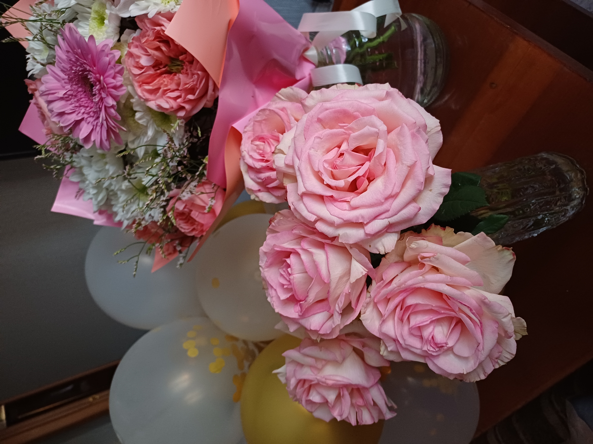 This is a close-up, eye-level shot of a flower arrangement and balloons. The subjects are a bouquet of pink and white flowers, primarily roses and gerbera daisies, and several balloons. The flowers are arranged in a pink paper wrapper. Some of the balloons are white and clear with gold confetti, while one is gold. The background consists of a dark wooden surface and a blurred glass vase with a white ribbon tied around its neck. The overall impression is festive and celebratory, suggesting an event like a birthday or anniversary. The lighting appears to be artificial, given the even illumination and lack of harsh shadows. There is no visible text in the image.