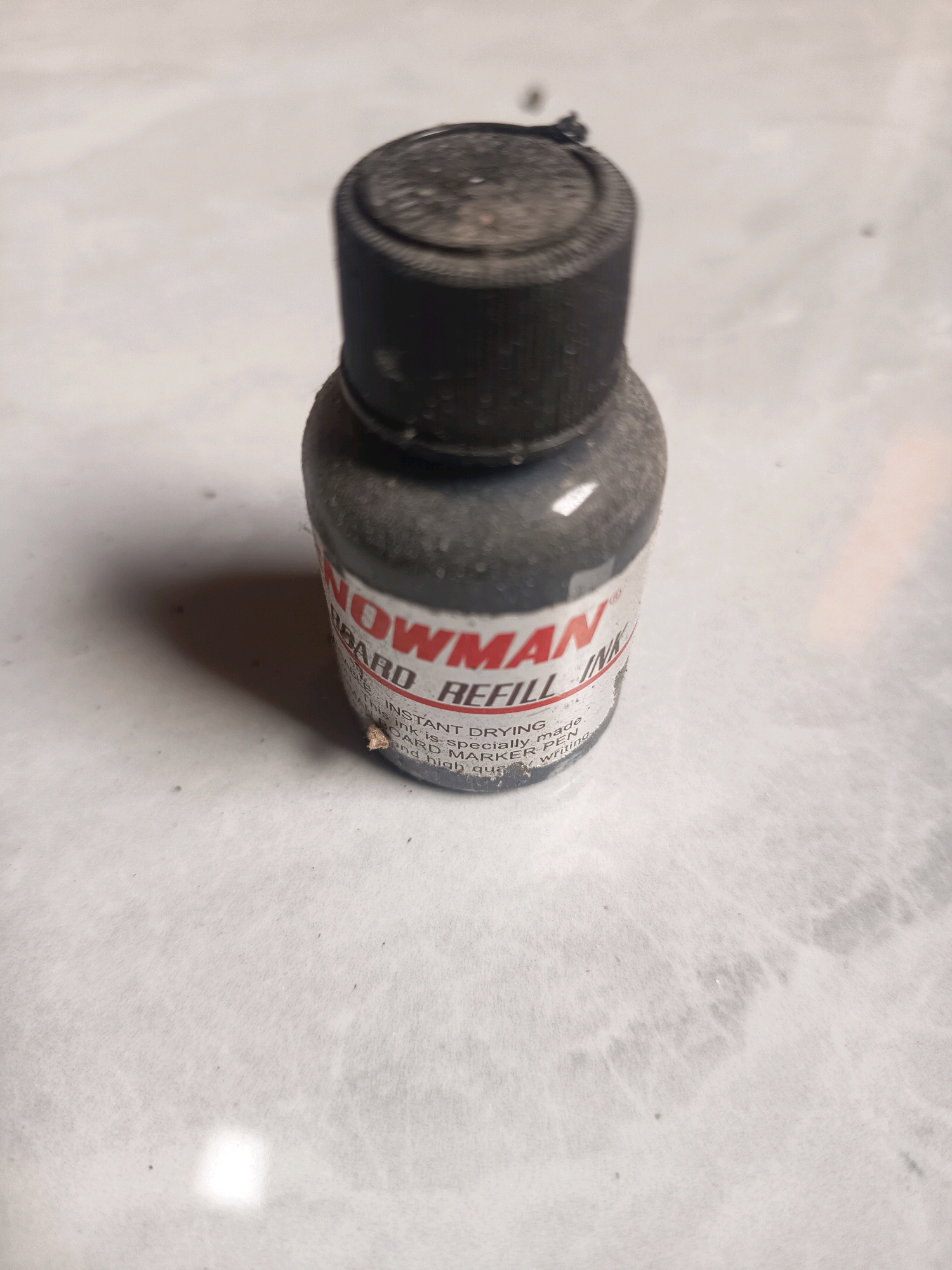 An overhead shot shows a small, dusty, black plastic bottle of ink refill. The label on the bottle is mostly red and white and reads "SNOWMAN BOARD REFILL INK". The bottle is placed on a light-colored, textured surface that appears to be a table or countertop, possibly in a home or office setting. There is no indication of location in Sidorejo, Indonesia.