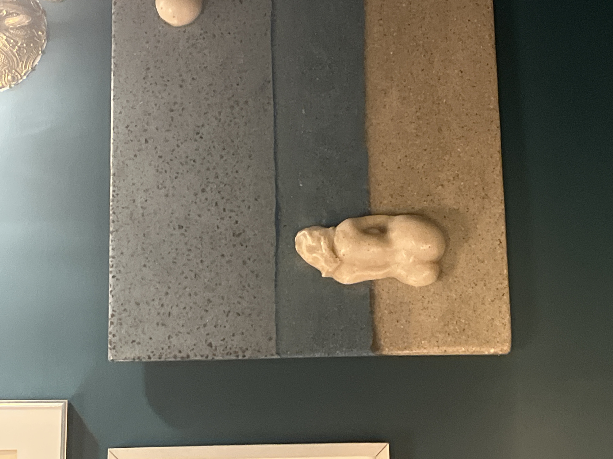 The image shows a piece of artwork mounted on a dark green wall. The artwork is a square panel with three vertical sections of varying textures and colors: speckled gray, matte dark gray, and speckled beige. Attached to the panel are two small, blob-like, beige-colored sculptures, one near the top of the gray section and one overlapping the transition between the dark gray and beige sections. The artwork is positioned above two white picture frames, only partially visible in the bottom left of the frame. The location, Lisbon, Portugal, is not discernible from the visual elements present in the photograph.