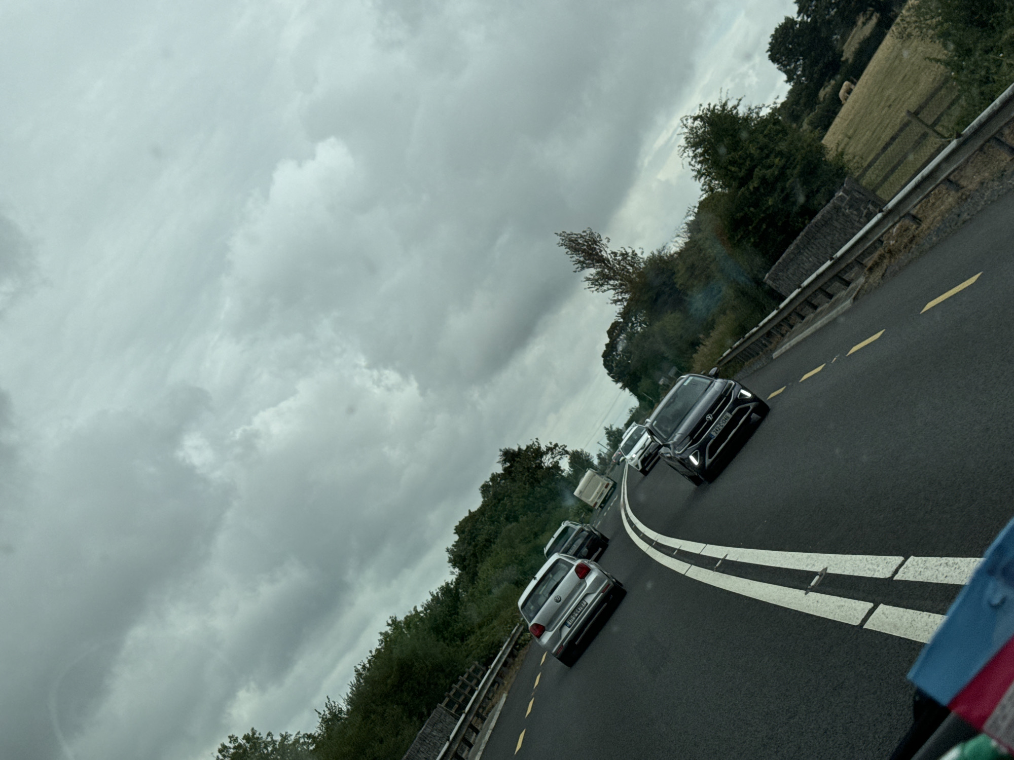 A traffic jam is depicted on a road in Scartnamore, Ireland. The overcast sky is filled with grey clouds. Several cars, including a silver Volkswagen hatchback, a black Volkswagen SUV, and a white van, are stopped on the asphalt road, which is marked with white and yellow lines. Trees and hedges line the side of the road, and in the distance, a field with a fence is visible. The perspective suggests the photo was taken from inside another vehicle.