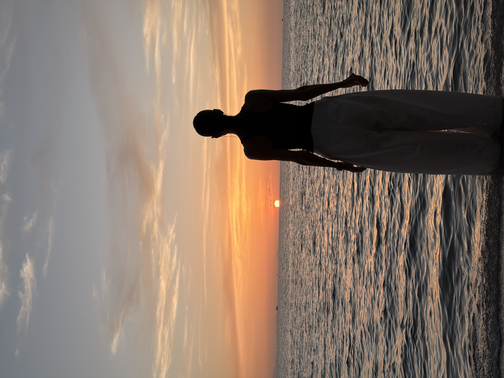A person stands on a beach at sunset, facing the ocean. The sky is a gradient of orange, pink, and blue, with wispy clouds. The sun, a bright orange orb, is setting on the horizon, casting a warm glow on the water. The waves are small and gentle, lapping at the shore. The person is silhouetted against the sunset, wearing a dark tank top and white, wide-leg pants. The person appears to be looking out at the ocean, perhaps contemplating the beauty of the scene. The location is the beach in Ko Samui District, Thailand.