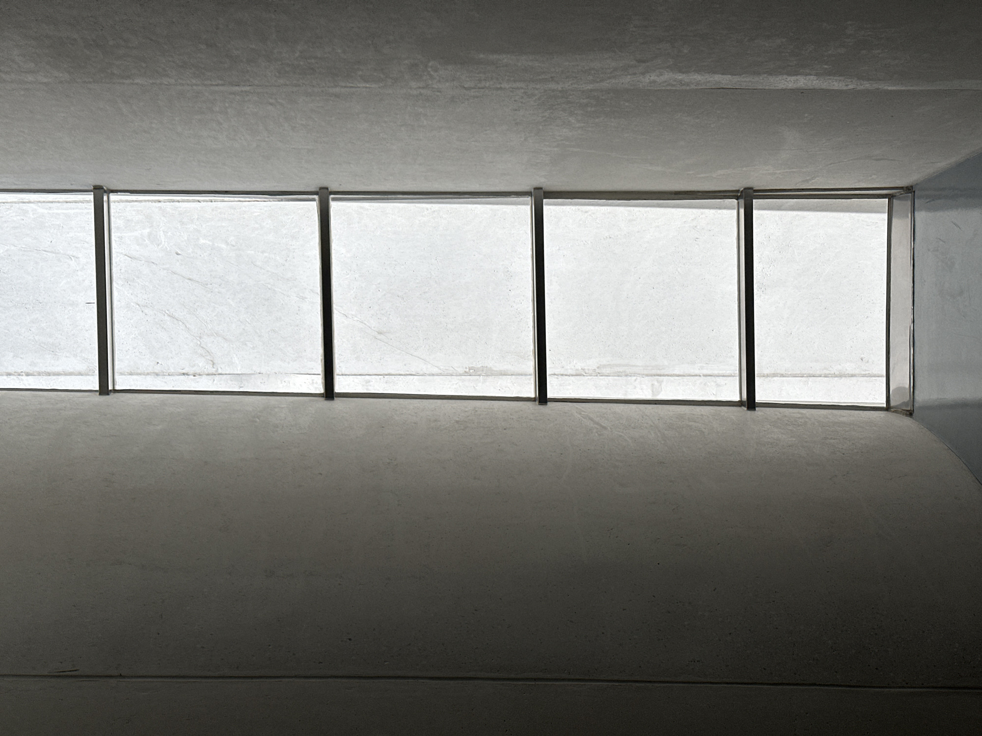 The image displays an architectural interior dominated by concrete surfaces and natural light filtering through a series of overhead openings. The perspective is looking forward and slightly upward within a structure that appears to be a tunnel, underpass, or a modern corridor.
In the upper half of the frame, a grey, textured concrete ceiling or upper wall is visible. Below this, a horizontal band features five distinct rectangular sections, likely windows or translucent skylights, framed by dark, vertical supports. These sections are brightly lit and appear to be made of a textured, light-colored material such as frosted glass or a similar translucent panel, showing subtle patterns and variations.
The lower half of the image presents a dark grey, textured concrete floor or ground surface, heavily shadowed and appearing flat. Towards the right side, the concrete surface suggests a gentle curve where it meets a wall, indicating a corner or a rounded architectural element. The overall lighting creates a high-contrast scene, with the bright light from the openings illuminating the dark, unadorned concrete interior.
No people, specific objects beyond the architectural elements, or discernible activities are present. The scene conveys a sense of starkness, minimalism, and quiet. While the exact time of day is not stated, the natural light suggests it is daytime. The location provided is San Bernardino Tlaxcalancingo, Mexico. There is no visible text in the image.