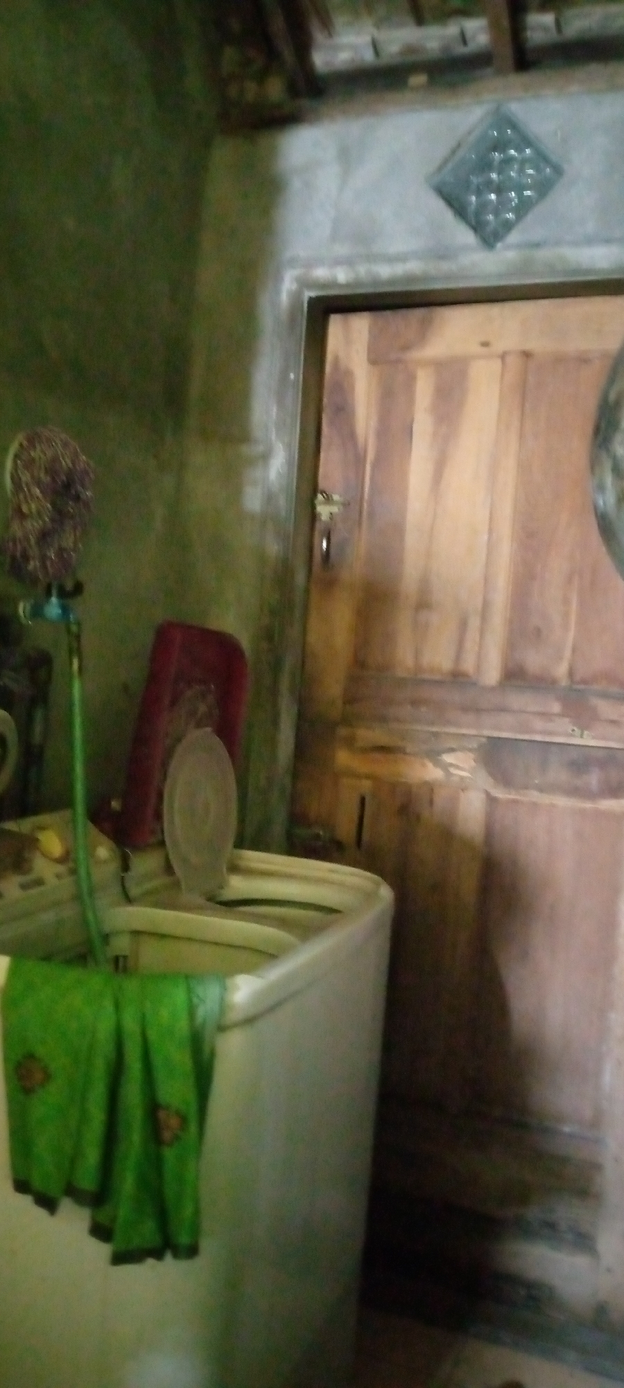 The image depicts an indoor scene, likely a laundry area within a residential dwelling in Sumberwono, Indonesia. In the foreground, a top-loading washing machine is visible, partially covered by a folded green cloth with a patterned border. To the left of the washing machine, a green hose is connected to a blue faucet, which has a brown mop head attached. Behind the washing machine, a red rectangular object, possibly a basket or container, is placed.
The background features a wooden door with vertical panels and a simple metal lock. Above the door, a diamond-shaped decorative element made of textured glass or similar material is embedded in the wall. The walls are painted in muted tones, with a greenish hue on the left and a gray concrete finish on the right. The ceiling appears to be constructed with wooden beams and planks. The overall impression is of a functional, lived-in space.