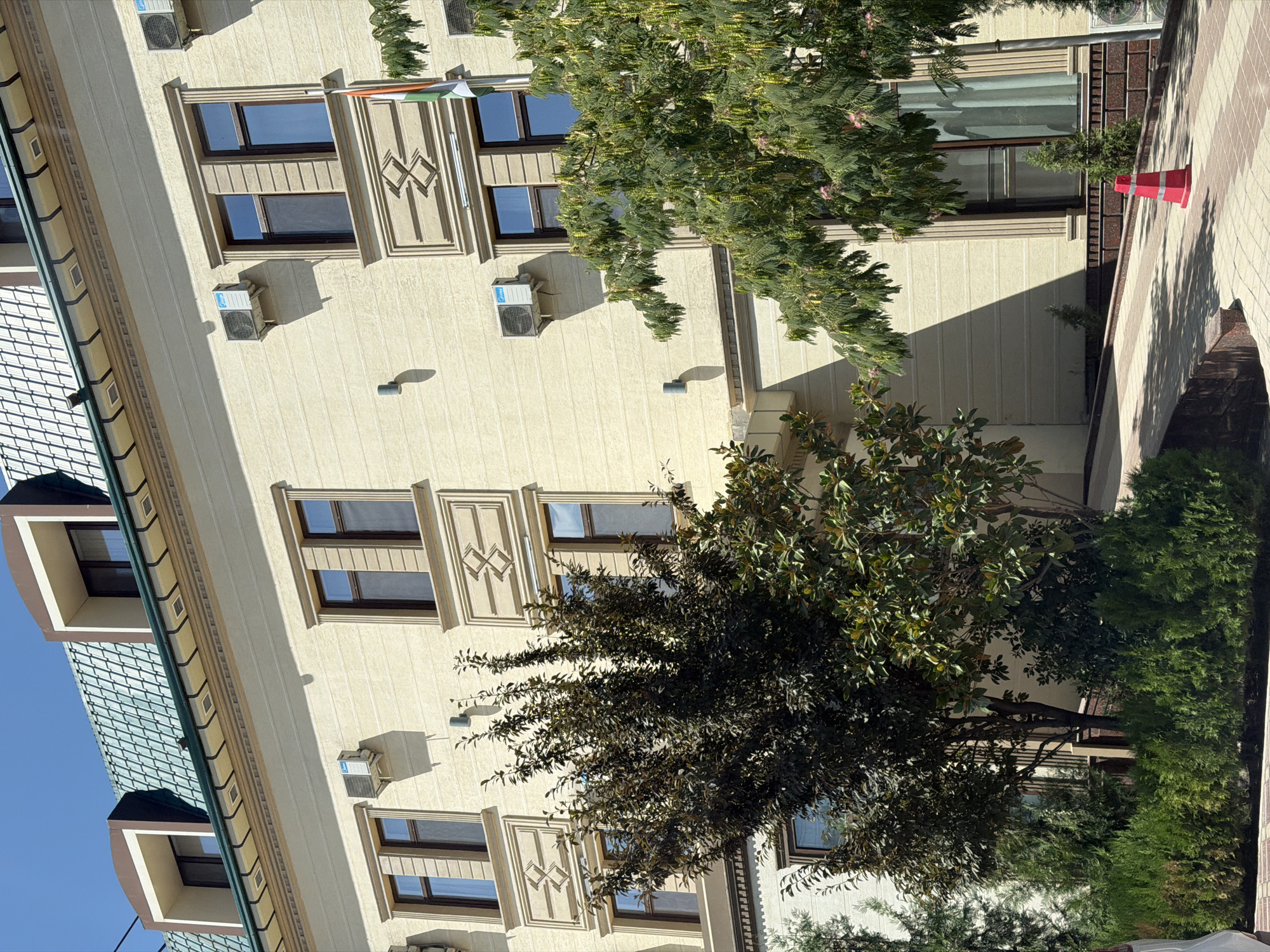 The image captures the exterior of a modern building with a beige facade, characterized by multiple windows, air conditioning units, and decorative stone elements above some of the windows. The roof features a distinctive green, tiled, mansard-style design with dormer windows. In the foreground, lush green trees and bushes are visible, casting shadows on a paved walkway that leads up to the building entrance. A red traffic cone stands on the walkway. The building appears to be located in a city, with a blue sky visible above. The presence of an Indian flag on the building suggests it may be an embassy or a business with ties to India, potentially located in the city of Tashkent, Uzbekistan.