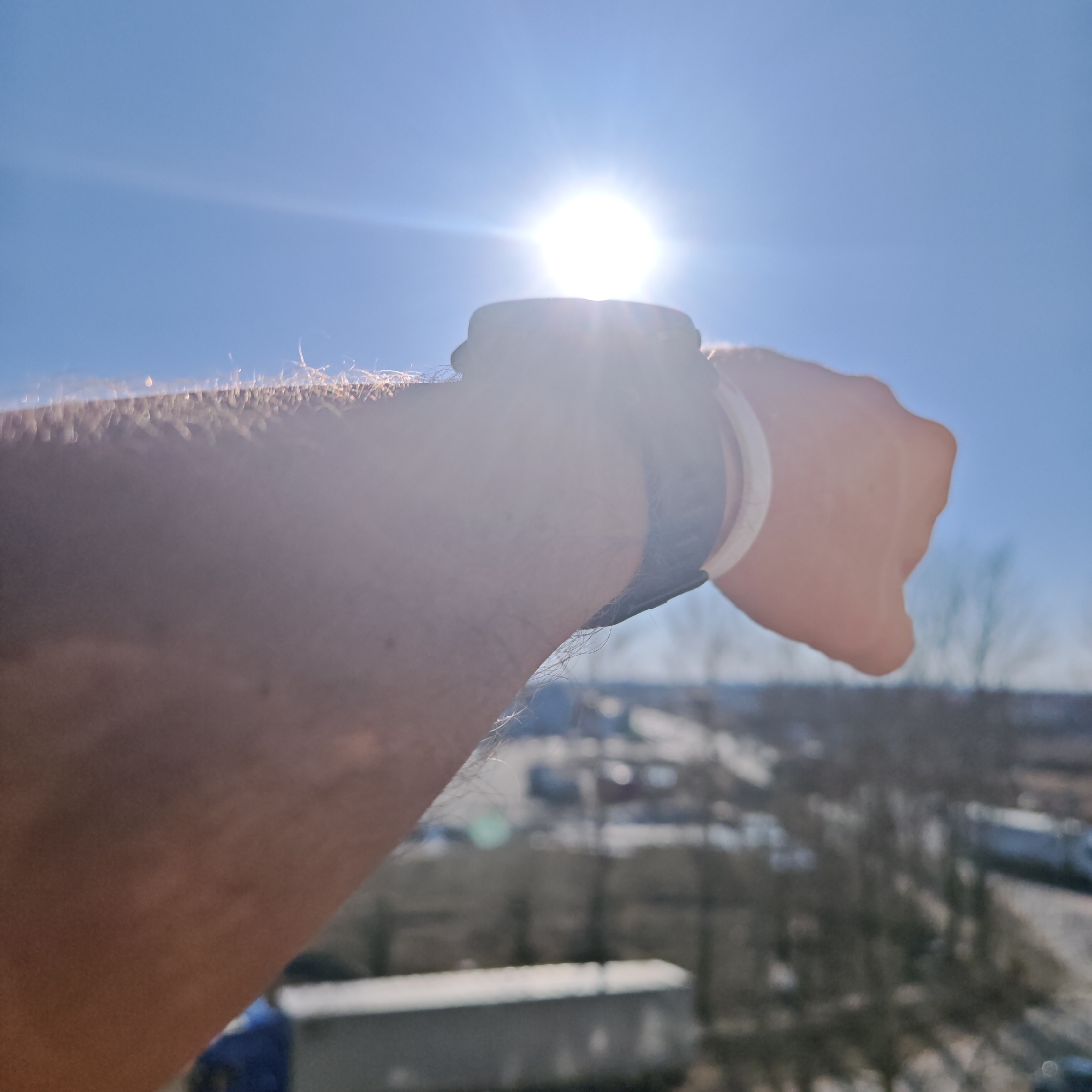 A person's arm is extended towards the bright sun in a blue sky. A wristwatch with a dark band and a light-colored band underneath is on their wrist. The background is blurry, showing what appears to be a cityscape with buildings, roads, and trees. The exact location in Rostov, Russia cannot be determined from the provided image.