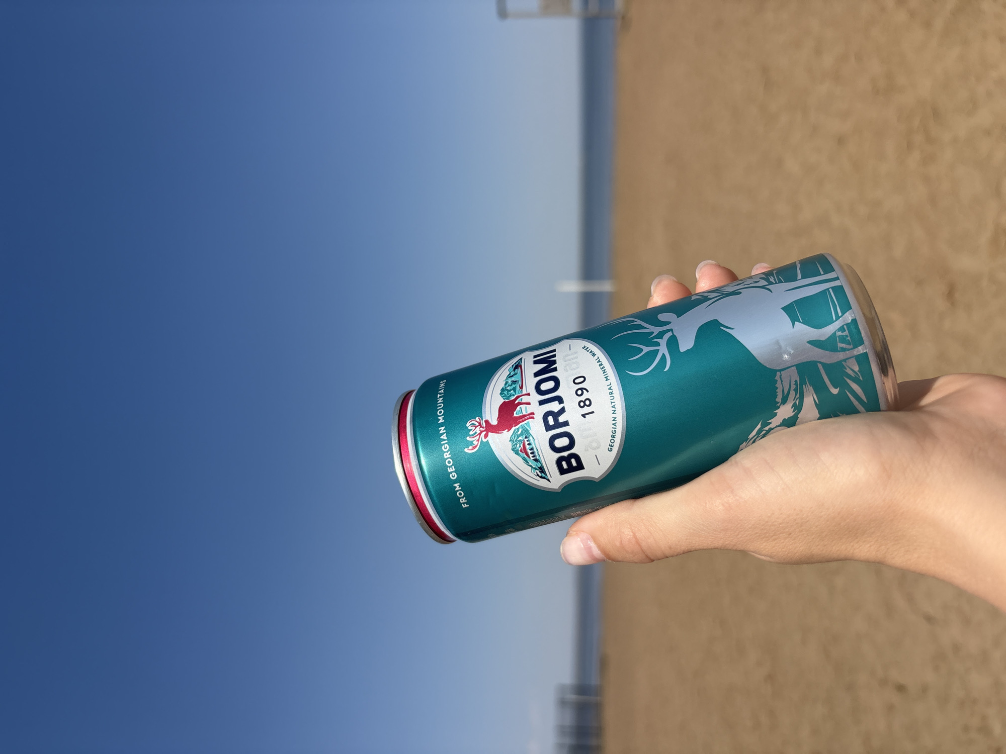 A hand holds a can of Borjomi mineral water against a backdrop of a blue sky and a sandy beach. The can is teal with a silver image of a deer. The red rims of the can are visible at both ends. The beach and a portion of a light-colored structure are visible in the background. The location is likely a beach area near Chaikovsky, Russia, although no landmarks in the image directly confirm the city.