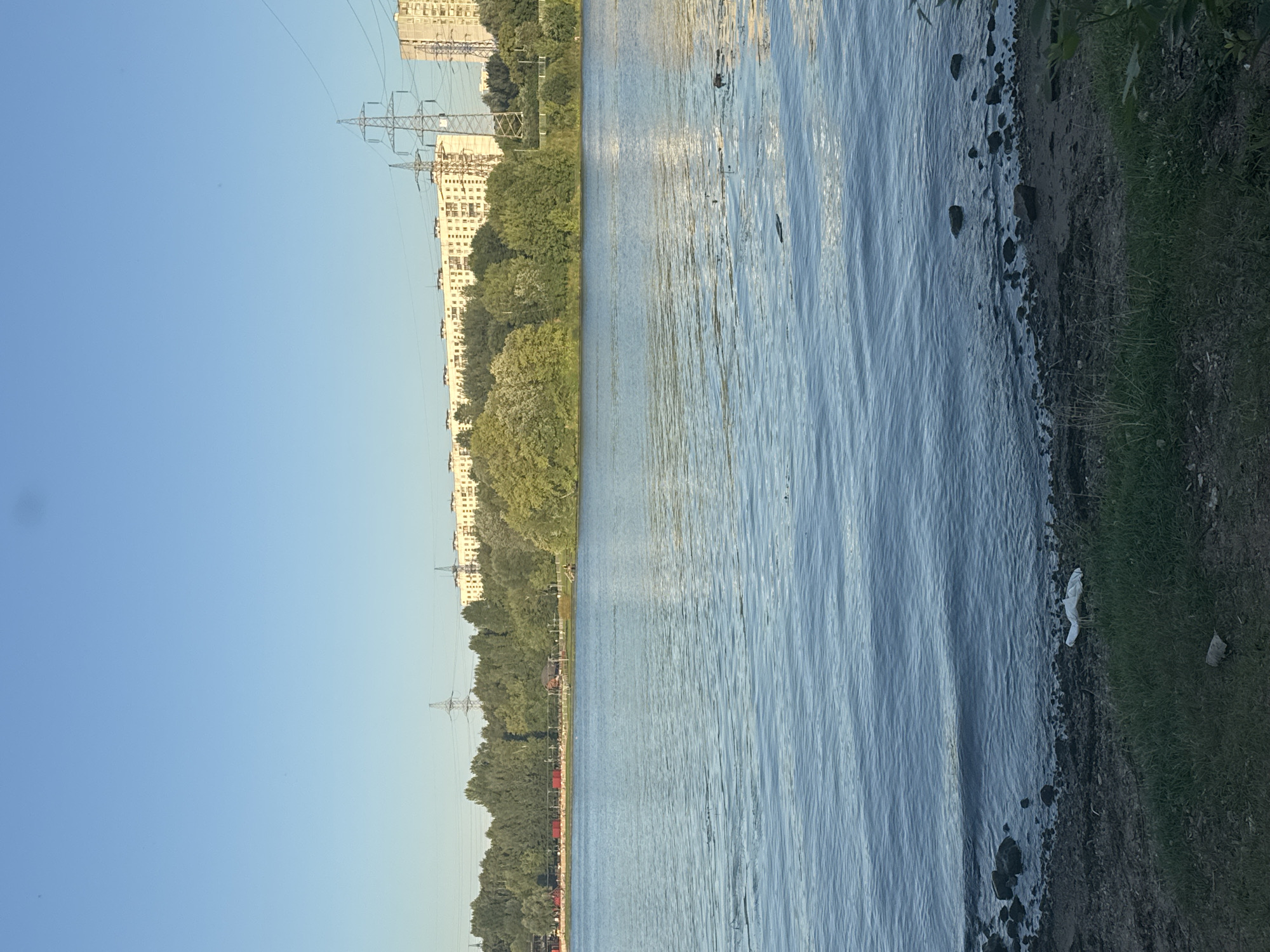 The image shows a body of water, likely a river or lake, with a shoreline in the foreground. The shoreline is rocky and has some vegetation, including grass and bushes. In the middle ground, there is a line of trees. In the background, there are several tall apartment buildings and some power lines. The sky is clear and blue. The scene appears to be a natural landscape within an urban environment. Specifically, this is a view of the Khimki Reservoir in Khimki, Russia, looking towards the residential areas of the city.