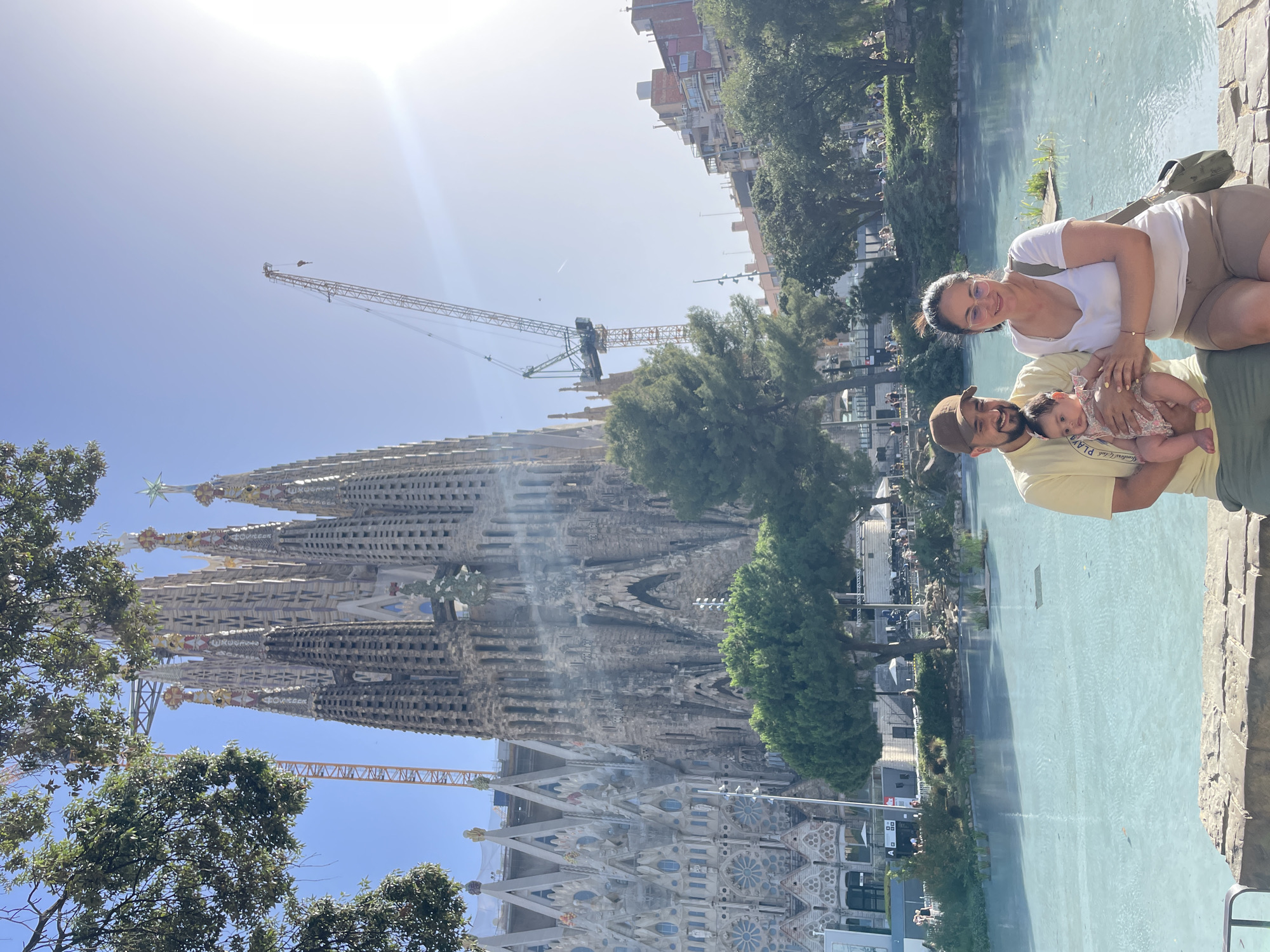 In Barcelona, Spain, a family of three is pictured in front of the Sagrada Familia. A man, woman, and baby are seated on a low stone wall. The man wears a tan baseball cap, a yellow t-shirt, and green shorts. He holds a baby wearing a floral-patterned dress. The woman wears glasses, a white t-shirt, and tan shorts. She has a green purse slung over her shoulder. The Sagrada Familia, a large unfinished basilica, dominates the background. Two cranes are visible behind the basilica, indicating ongoing construction. A bright blue body of water lies in front of the family, and green trees are scattered throughout the scene. The sun is shining brightly, creating a lens flare in the upper right corner of the image.