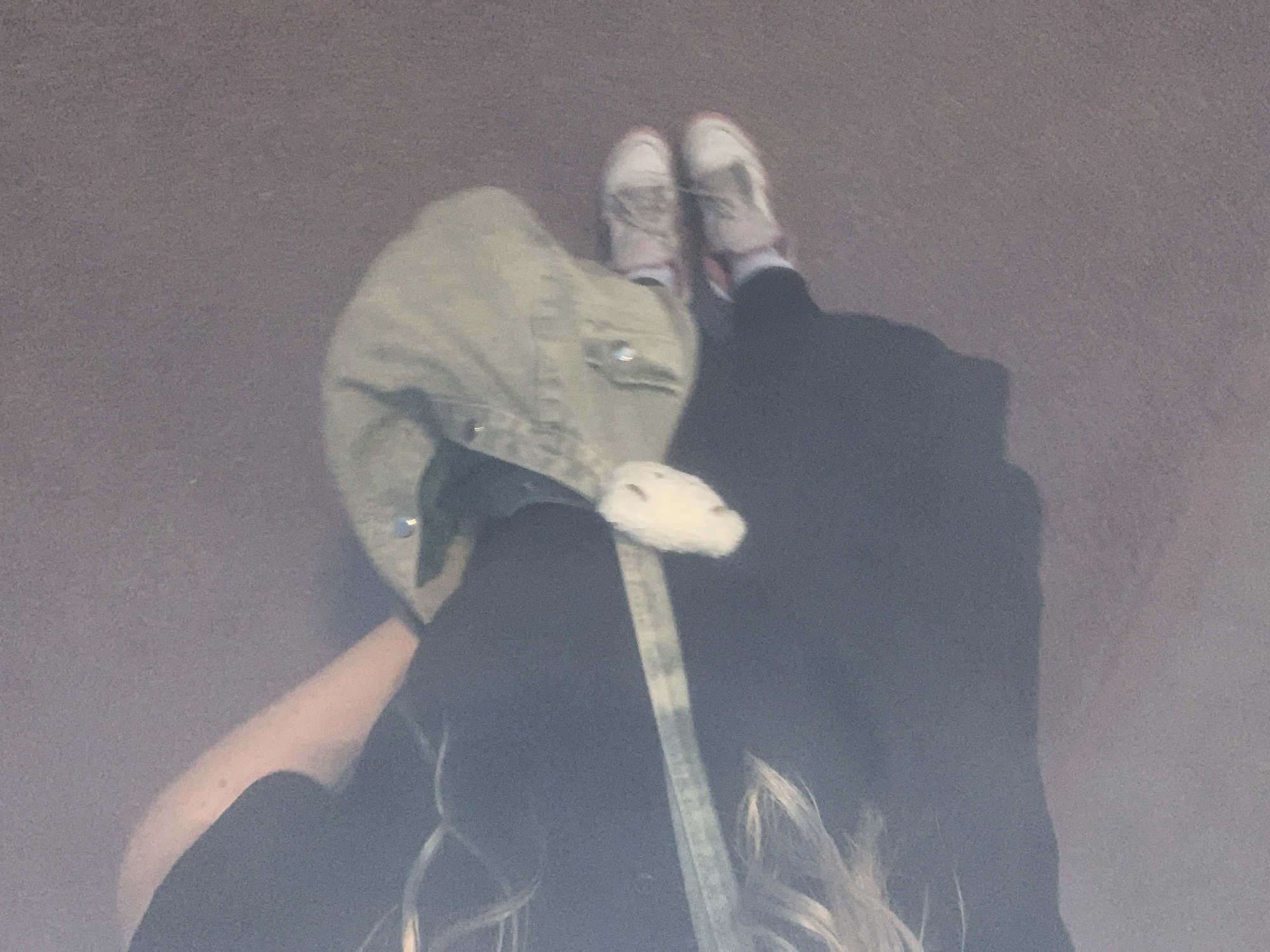 The image shows a person wearing a black outfit and sneakers, walking on an asphalt surface. The person is carrying a light-colored shoulder bag. The background is a blurry, grayish asphalt. The image appears to be taken outdoors, possibly in an urban or suburban area. There is no visible indication of the specific location within the city of Vsevolozhsk, Russia, or any landmarks. The lighting suggests it might be daytime, but the overall quality of the image is low, with significant noise and motion blur. The perspective is from above and slightly to the side, looking down at the person's legs and feet.