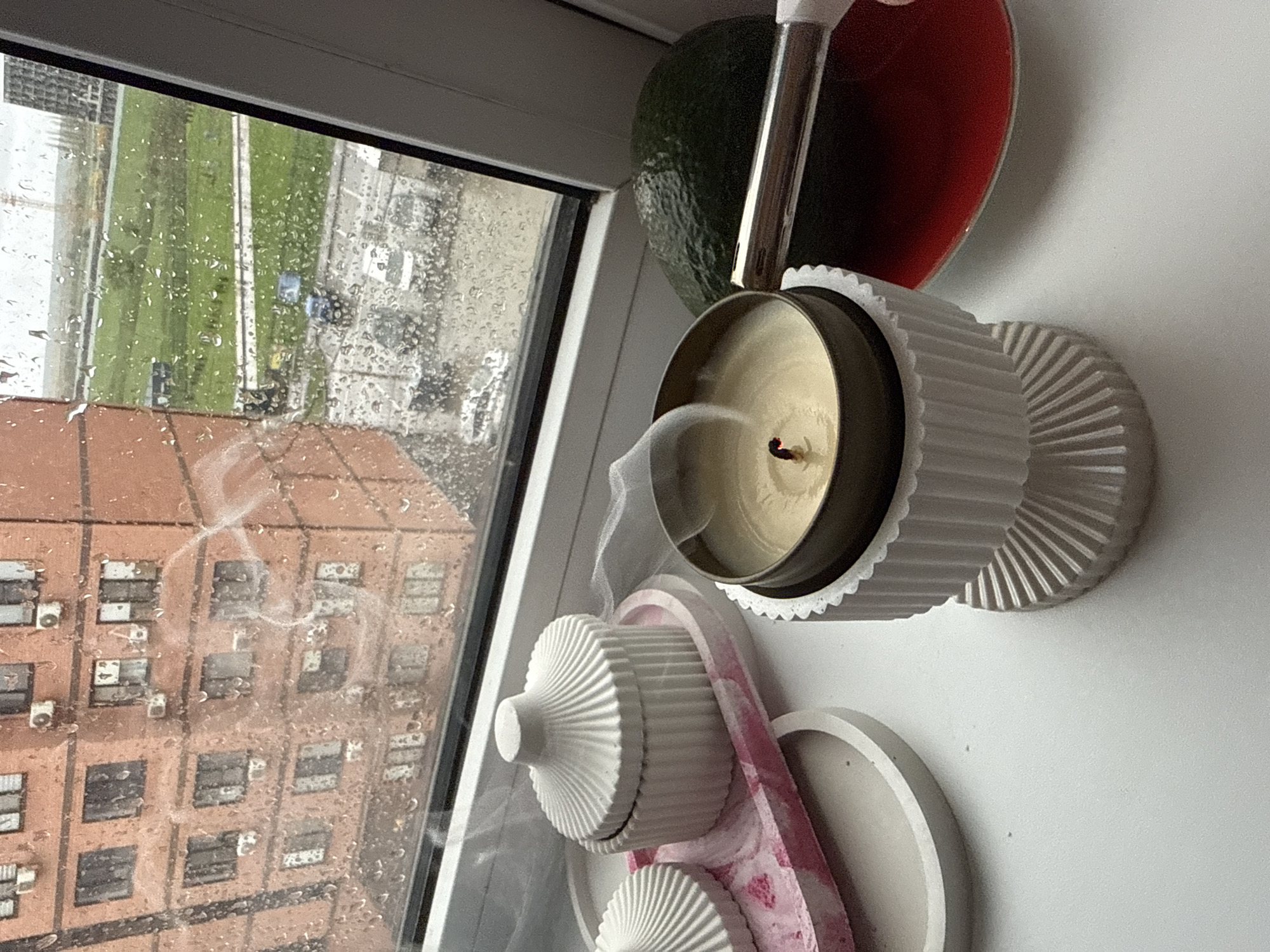 A candle is burning on a windowsill on a rainy day. The windowsill also has some other art objects including an avocado and a red shape. The building outside the window has rows of windows with air conditioners.