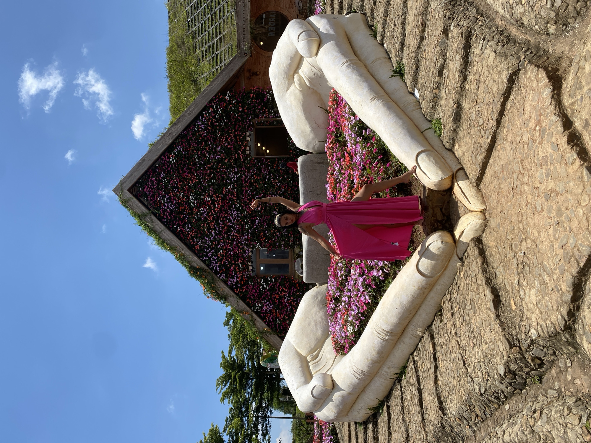 In the city of Cam Ly, Đà Lạt, Vietnam, a woman in a vibrant pink dress stands within a large, heart-shaped arrangement of white stone hands. The hands, sculpted to appear as if gently cupping the ground, frame a bed of pink and purple flowers. Behind this floral display, a house with a roof covered in greenery and flowers serves as a backdrop. The sky above is a clear blue with scattered white clouds. The ground leading up to the hands is paved with stones, creating a rustic pathway.