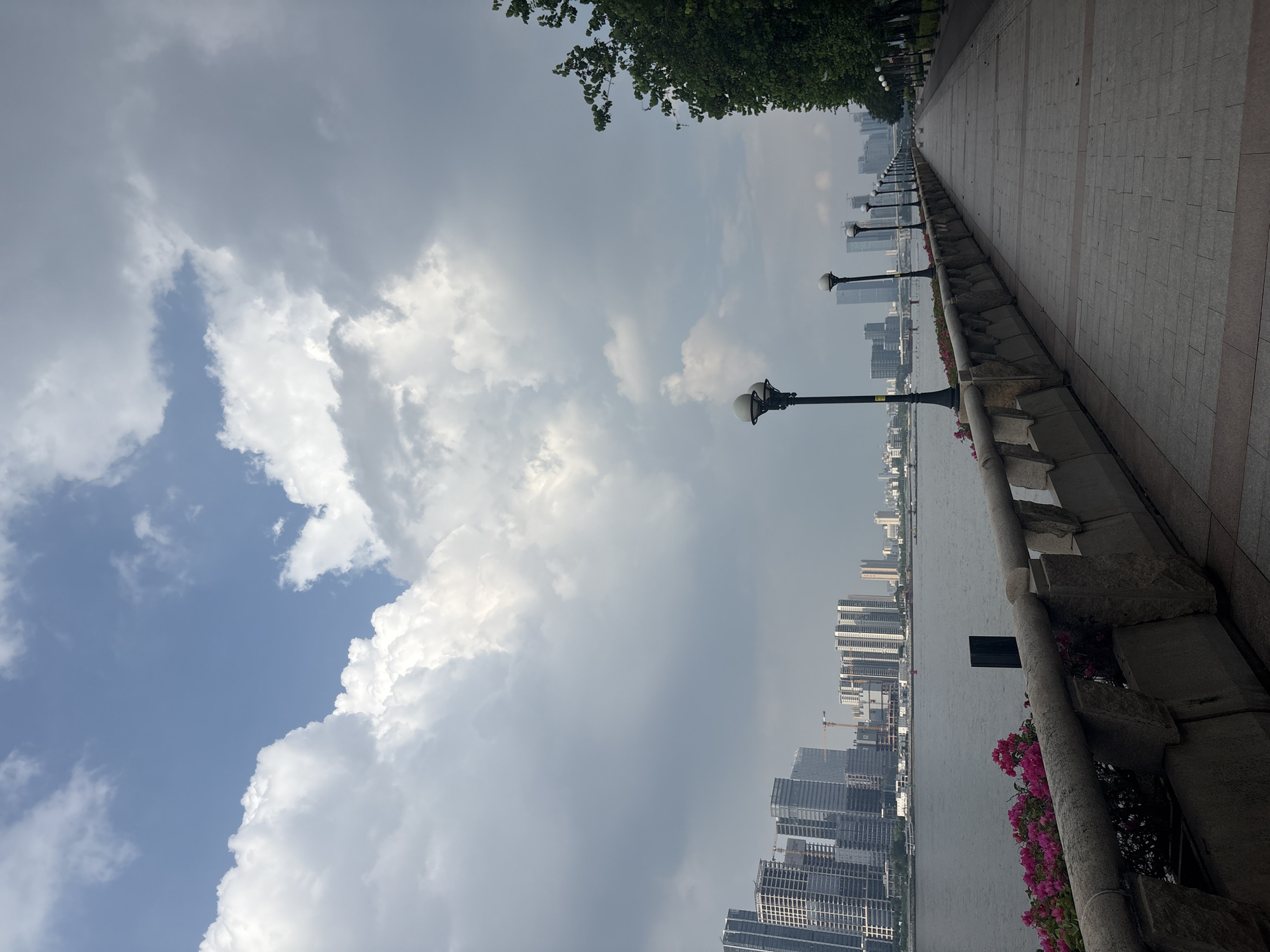 The image depicts a picturesque waterfront promenade in Haizhu District, China. The scene is dominated by a vast sky filled with dramatic, billowing clouds that hint at changing weather, possibly a storm approaching or just passed. The clouds cast shadows on the cityscape in the distance, creating a dynamic interplay of light and dark.
On the right side of the frame, a meticulously paved walkway stretches into the distance, lined with a stone railing. Several ornate lampposts with round globes are evenly spaced along the promenade, suggesting it is a public space designed for leisurely strolls, likely to be used at dusk or night. Beside the railing, vibrant pink flowers add a touch of color to the otherwise muted tones of the stone and pavement.
Across the tranquil body of water, a dense skyline of modern high-rise buildings rises, indicating an urban environment. The architecture is contemporary, with many glass-fronted towers. Construction cranes are visible among the buildings, suggesting ongoing development. The water appears calm, reflecting the overcast sky and the distant city.
The overall mood is serene yet powerful, with the dramatic sky and imposing city creating a sense of awe. The time of day is likely late afternoon or early evening, given the quality of light and the presence of the lampposts. The location cues – the specific style of buildings, the waterfront setting, and the known geographical context of Haizhu District – strongly anchor the scene. No text is visible in the image.