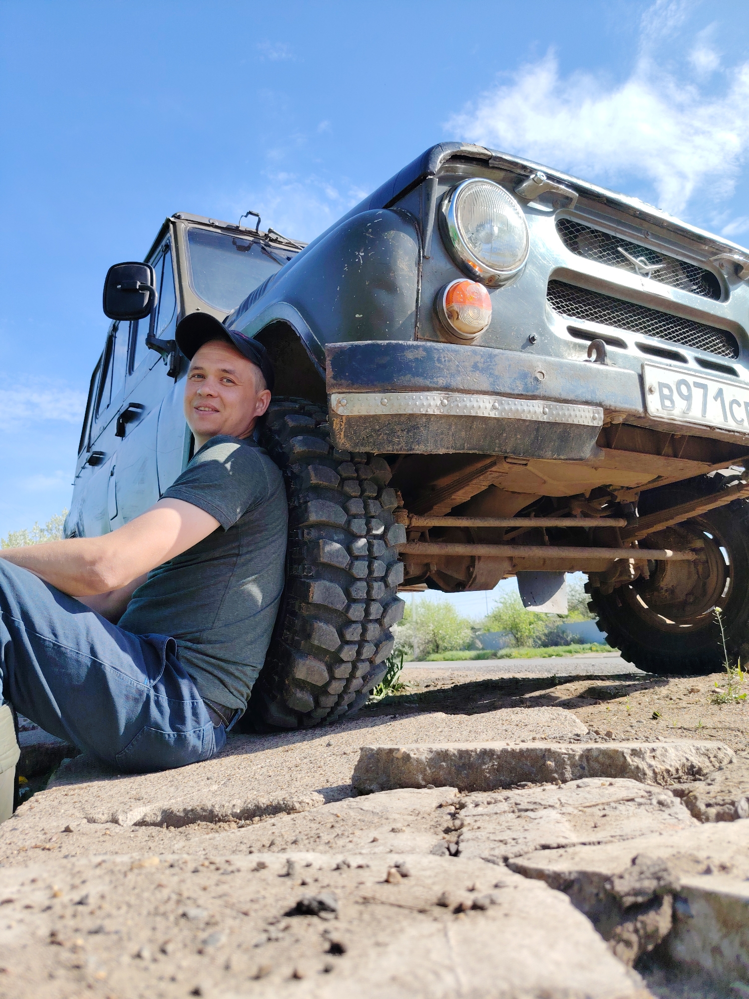 A man is sitting on cracked concrete near a green off-road vehicle in Chaikovsky, Russia. The man is wearing a black baseball cap, green t-shirt, and blue jeans. He is leaning against the front tire of the vehicle. The vehicle is an older model with a Russian license plate "B971CC". The scene is outdoors on a sunny day. A road and some green vegetation are visible in the background.