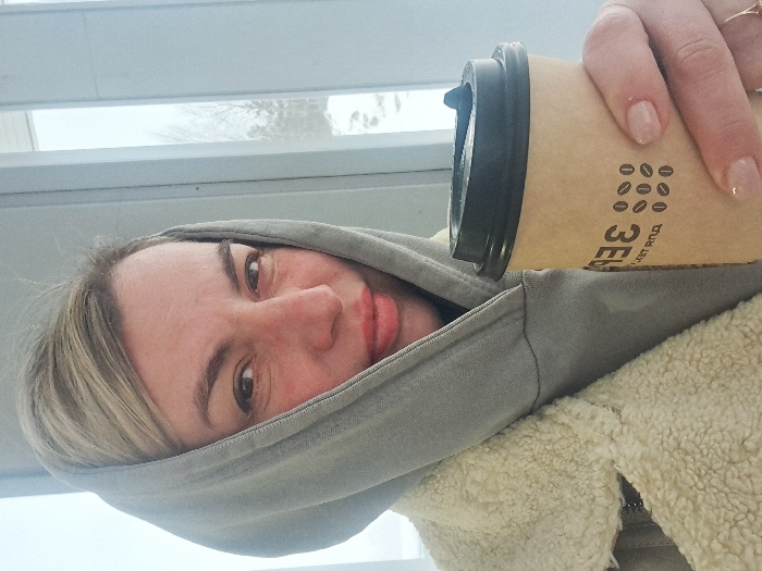 A woman with fair skin and blonde hair, wearing a gray hooded jacket, holds a brown paper coffee cup with a black lid. The cup has a coffee bean design. She is looking directly at the camera with a slight smile. A window with horizontal blinds is visible in the background. She is wearing a ring on her right ring finger. Her nails are painted a neutral shade. She has light-colored jacket underneath the hoodie.