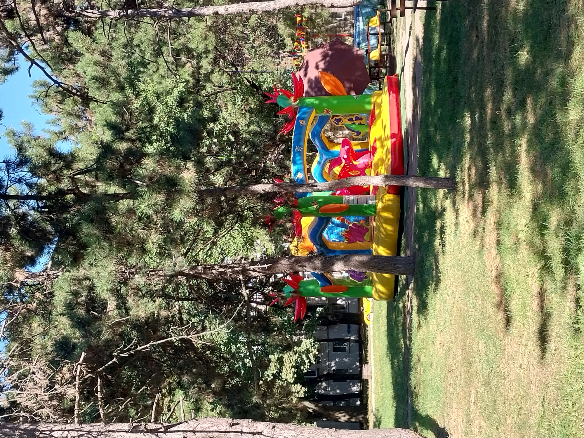 This is an outdoor, eye-level, slightly low-angle, full-shot photograph taken on a sunny day, likely in a park or recreational area within the city of Dnipro, Ukraine. The scene features a colorful inflatable playground structure, possibly a bounce house, positioned on a grassy area. The playground is predominantly yellow, red, blue, and green, with some red and green inflatable decorations resembling flowers or foliage on top. Inside the inflatable structure, dinosaur figures are visible.
In the background, trees with dense green foliage dominate the scenery, with some older, taller trees in the foreground casting shadows on the grass. Behind the playground and to the left, a multi-story building with windows can be seen, partially obscured by trees. A yellow toy car is parked on the grass to the left of the inflatable playground. Further to the right, a brown umbrella shades a picnic table with yellow benches. The overall environment suggests a public space designed for recreation, possibly a children's park or an event venue. The lighting indicates it is daytime, with the sun casting distinct shadows. There are no people clearly visible in the image.