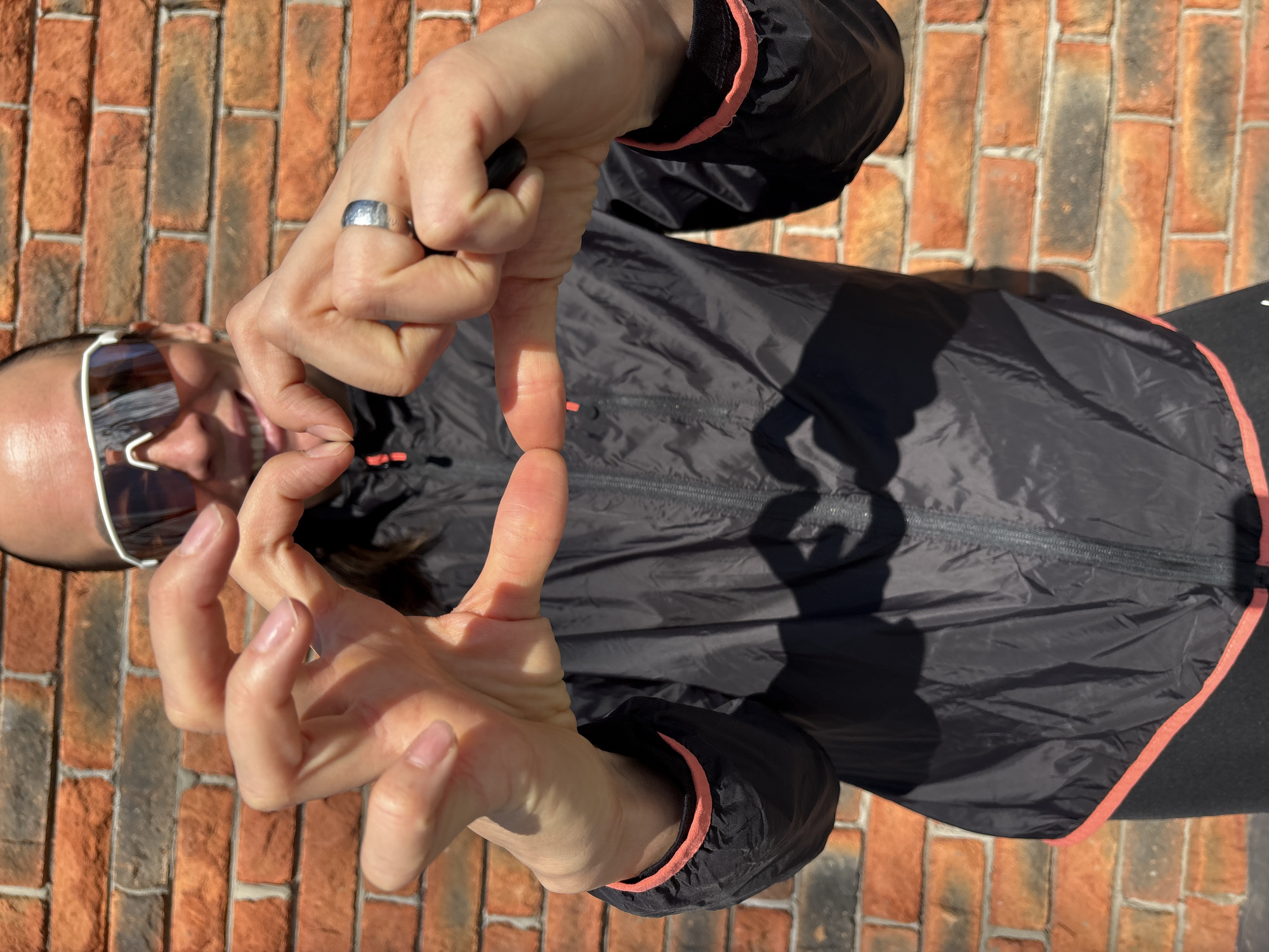 A high-angle outdoor shot shows a person standing in front of a brick wall. The person is fair-skinned with light-colored hair. They are wearing white-framed sunglasses and a dark jacket with a coral pink trim. The person is making a square with their fingers and looking through the opening. A wide silver ring is visible on the person's left hand. The background is a brick wall. Based on available information, the location of this scene in the city of Chaikovsky, Russia, cannot be determined.