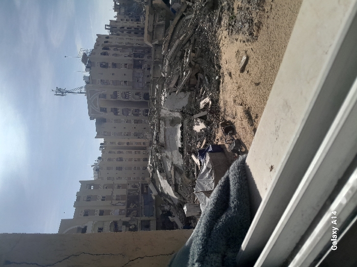 The image depicts a heavily damaged urban landscape in Khan Yunis, Palestinian Territory, under a bright, partly cloudy sky. The perspective is from a window or opening, with a cracked light-brown wall section on the left and a gray, textured fabric resting on a light-colored windowsill in the foreground.
The midground reveals extensive destruction, dominated by large piles of concrete rubble, twisted rebar, and debris scattered across sandy, uneven ground. Amidst this rubble, a blue tarp covers an object or forms part of a small, rudimentary structure, with another light-colored, possibly tent-like structure nearby.
In the background, a row of multi-story buildings, approximately five to seven stories high, extends horizontally. These structures, characterized by multiple windows and balconies, exhibit significant damage, including missing walls, blown-out windows, and scarred facades, suggesting extensive structural compromise. One prominent building in the center-right features a distinct arched architectural detail near its roofline. A communications tower, equipped with multiple antennas, is mounted on the roof of this central building, alongside other smaller rooftop structures and antennas. No individuals are visible in the scene.