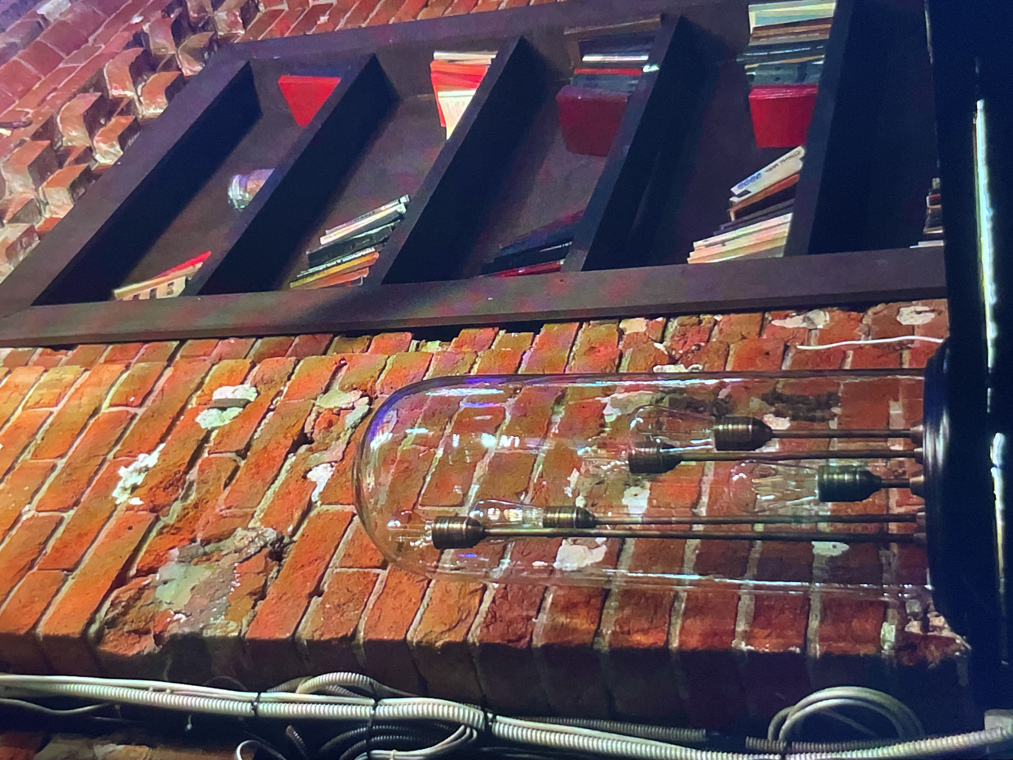 The image depicts a detailed view of an interior wall, seemingly within a building located in Unknown, Unknown. The wall is constructed of red brick, exhibiting signs of wear and age, with patches of missing mortar and discoloration. Affixed to the wall is a large, transparent glass tube containing several light bulbs attached to metal rods. Below the glass tube, a bundle of gray and white electrical cables and conduits runs horizontally along the wall. Above the wall is a dark wooden structure that resembles a shelf or display case, holding a collection of books and objects of various colors, including red. The shelf is angled, and its contents are not clearly visible, suggesting a display of objects stored or exhibited on shelves. The setting appears to be a building with a rustic or industrial aesthetic.