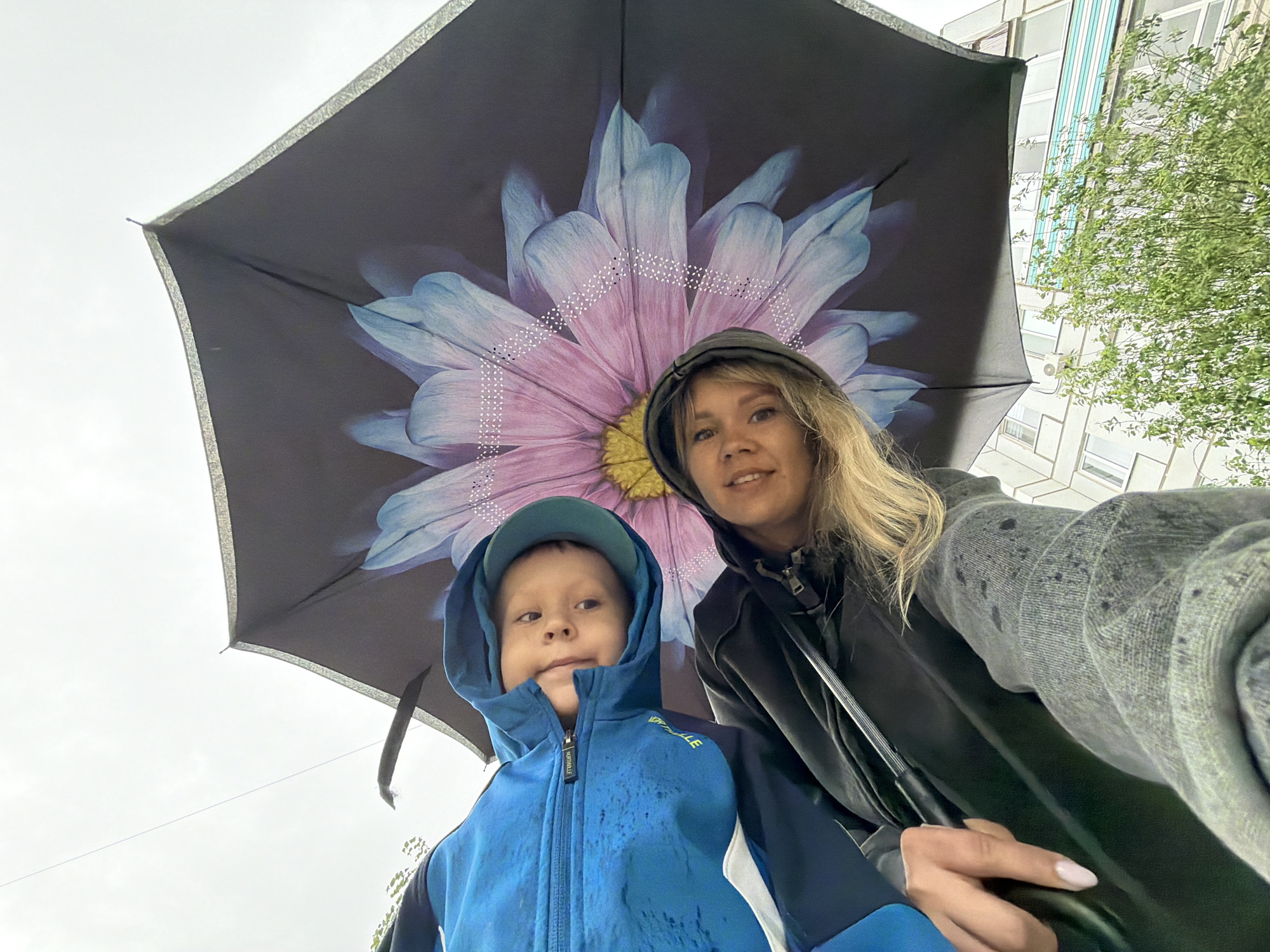 A woman and a child stand under an umbrella with a large floral design, possibly in a city that is Chaikovsky, Russia. The woman is wearing a dark hooded jacket and the child is wearing a blue hooded jacket and a cap. The background includes a building with light-colored walls and windows, and some green foliage. The sky is overcast. The woman is holding the umbrella.