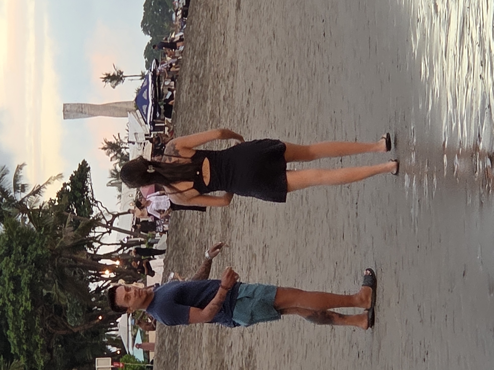 In North Kuta, Indonesia, on a beach, a man and a woman are standing near the water's edge. The man, facing the camera, wears a dark blue t-shirt and teal shorts, with intricate tattoos visible on his legs and arms. He is looking towards the woman. The woman, with her back to the camera, is wearing a black dress with a cutout detail on the back. She is looking away from the man, towards the ocean. The beach is sandy, and the water is shallow and reflective. In the background, palm trees line the shore, and other beachgoers and umbrellas are visible. A tall, modern concrete structure stands in the distance. The sky above is cloudy, with soft hues of pink and orange suggesting it is either dawn or dusk.