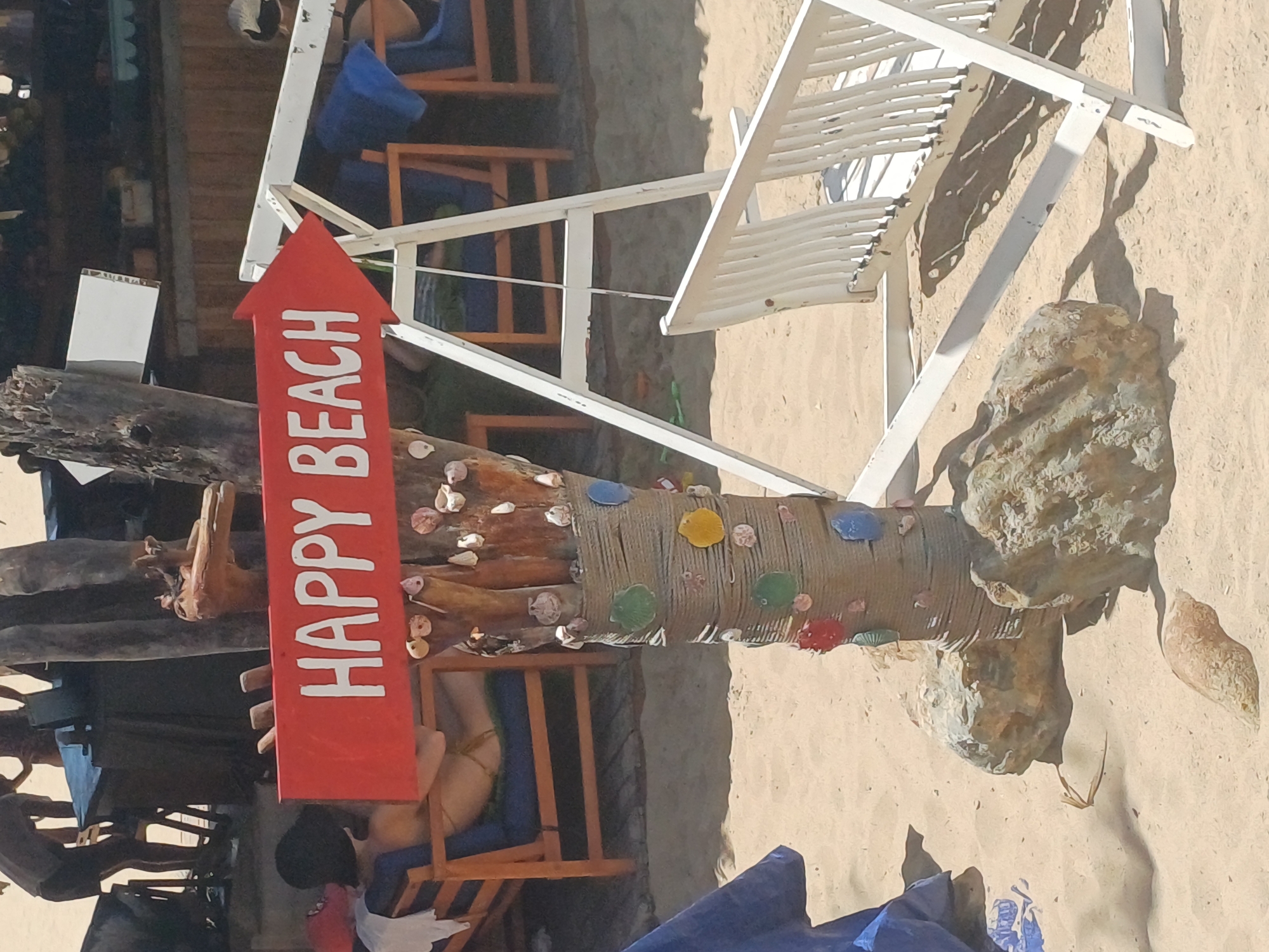 A red arrow-shaped sign with white lettering reading "HAPPY BEACH" is mounted on a wooden pole decorated with shells and twine. The pole is situated on a sandy beach. Behind the pole, a white A-frame structure supports a white wooden swing seat, partially visible. In the background, there are several people and beach chairs. One person is sitting in a wooden lounge chair, wearing a black hat and a bikini. Another person is standing in the background near a table. The environment suggests a tropical beach setting in Nha Trang, Vietnam.