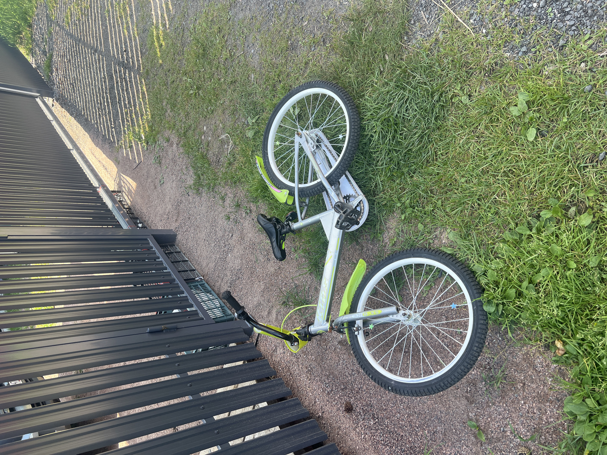 A children's bicycle with a silver frame and lime green accents is lying on its side on a patch of grass next to a gravel path and a dark grey metal fence. The bicycle is a BMX-style bike with knobby tires and a black seat. The front wheel is pointing towards the camera and is slightly turned. The rear wheel is visible behind the frame. The fence appears to be part of a property boundary. The scene is outdoors and appears to be in a residential area, possibly a dacha or garden community, given the presence of the fence and the natural vegetation. The lighting suggests it is daytime with sunlight casting shadows from the fence onto the ground.