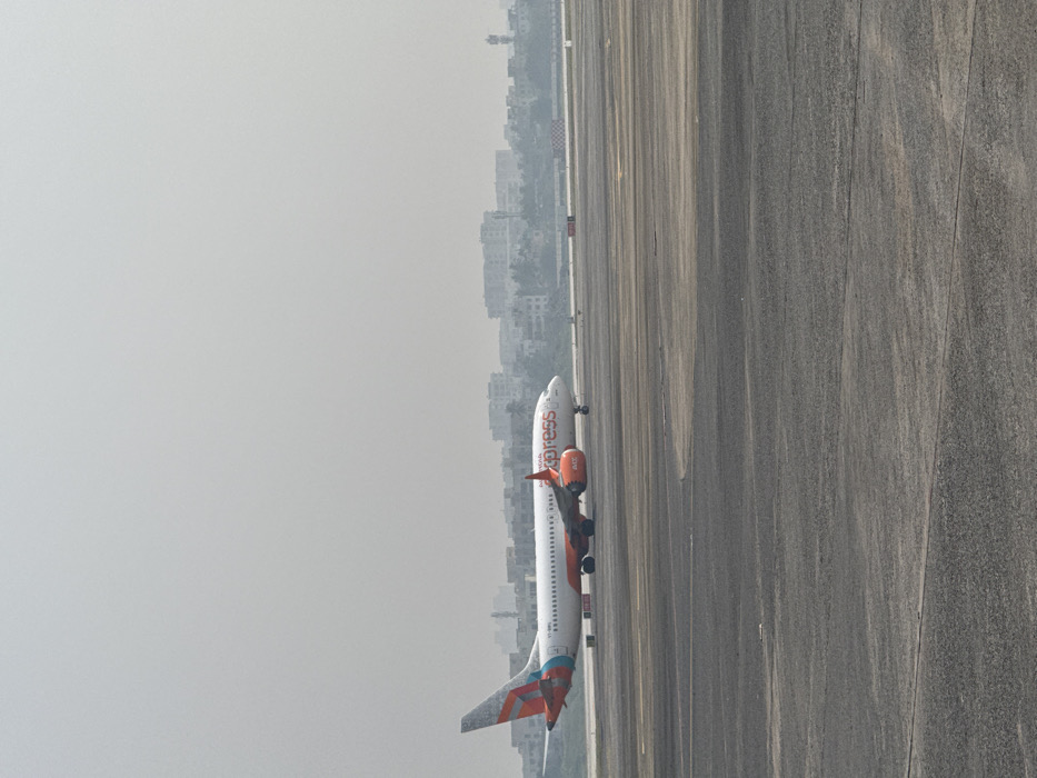 An airplane, predominantly white with orange accents, is positioned on a gray runway. The aircraft is viewed from a slightly elevated angle, showing its side profile. In the background, a hazy skyline of buildings is visible, suggesting an urban area. The overall atmosphere is overcast, casting a soft light on the scene. The runway lines converge toward the distance.