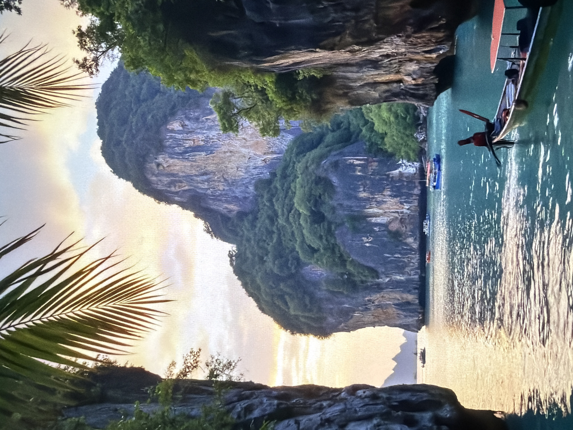 The image depicts a breathtaking tropical landscape, likely at sunrise or sunset, characterized by towering limestone karsts rising dramatically from calm, turquoise waters. The scene is bathed in warm, soft light, casting hues of yellow, orange, and soft blue across the sky and reflecting brightly on the water's surface.
On the right side of the frame, a traditional longtail boat with a distinctive red canopy is prominently featured on the water. A person wearing a red shirt is visible near the front of the boat, possibly preparing to operate it or disembark. Further in the mid-ground, several other smaller boats are scattered across the water, surrounded by the majestic rock formations.
The left side of the image is dominated by a massive, rugged cliff face, densely covered in lush green vegetation, extending from the foreground into the mid-ground. Another landmass with greenery is visible in the bottom left. A large, graceful palm frond partially frames the upper left corner, enhancing the tropical ambiance. The water displays shimmering reflections from the soft light, creating a peaceful and picturesque setting. The overall impression is one of serene natural beauty and exploration, typical of coastal regions in Southeast Asia. No text is visible in the image.