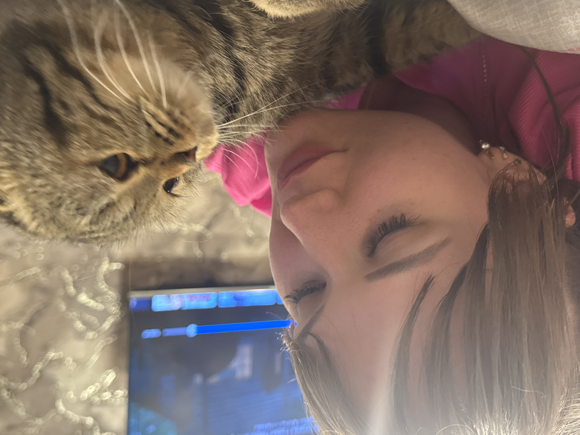 This close-up image captures an intimate, tender moment between a tabby cat and a person, likely a woman, indoors in Chaikovsky, Russia. The image is oriented vertically, showing the subjects at an angle.
The cat, with its distinctive brown and grey striped fur and long, prominent white whiskers, is positioned on the left side of the frame. Its face is turned towards the person, with one alert, amber-colored eye clearly visible as it appears to sniff or nuzzle the person's face.
The person's face is seen in profile on the right, from just above the eyebrow down to the chin. Her eyes are closed or nearly closed, and her lips are slightly parted. She has long, dark hair, with some strands softly framing her face, and is wearing a vibrant pink garment, possibly a top or a blanket draped around her. A small, white earring is visible on her earlobe. The close interaction suggests a bond of affection and companionship.
In the background, a digital screen, possibly a laptop or tablet, is visible, displaying a user interface with bright blue and white graphical elements, including what appears to be a progress or volume slider. To the left of the screen, a light-colored, textured fabric, resembling bedding or a blanket, is out of focus. The artificial lighting suggests an indoor setting, likely evening or night, contributing to a cozy and relaxed atmosphere. No discernible text is visible on the screen.