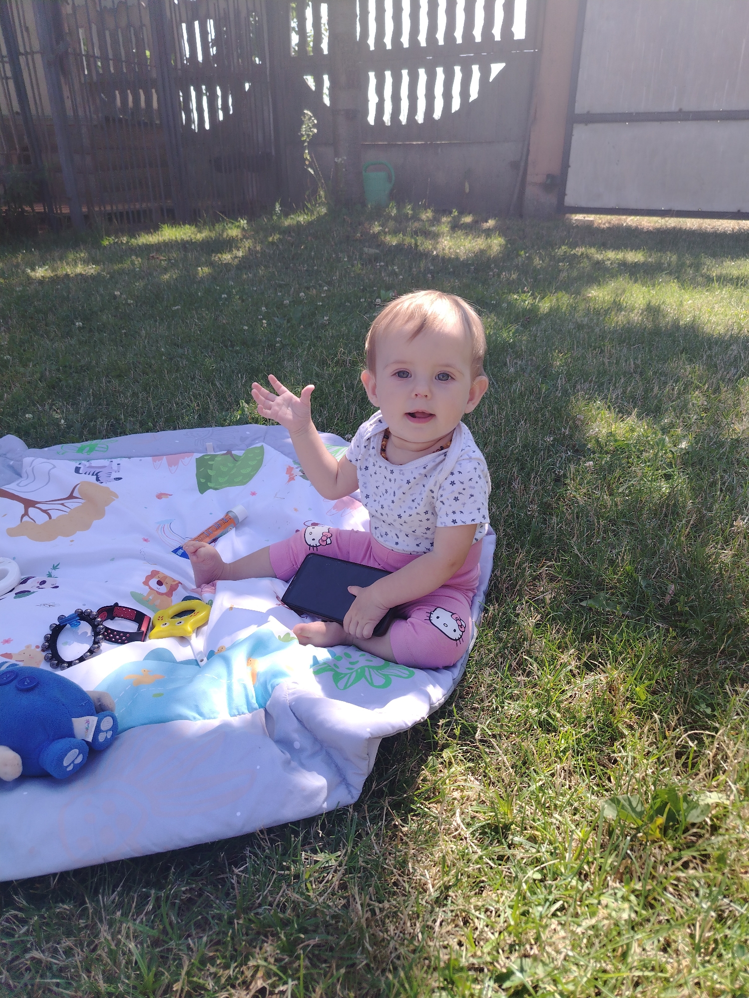 A baby sits on a blanket in a grassy yard. The baby is wearing a white onesie with a floral pattern and pink pants with a Hello Kitty graphic. The baby is holding a black smartphone. There are several toys on the blanket, including a blue stuffed animal, a yellow toy, and a beaded bracelet. In the background, there is a wooden fence and a green watering can. The scene appears to be a sunny day.
