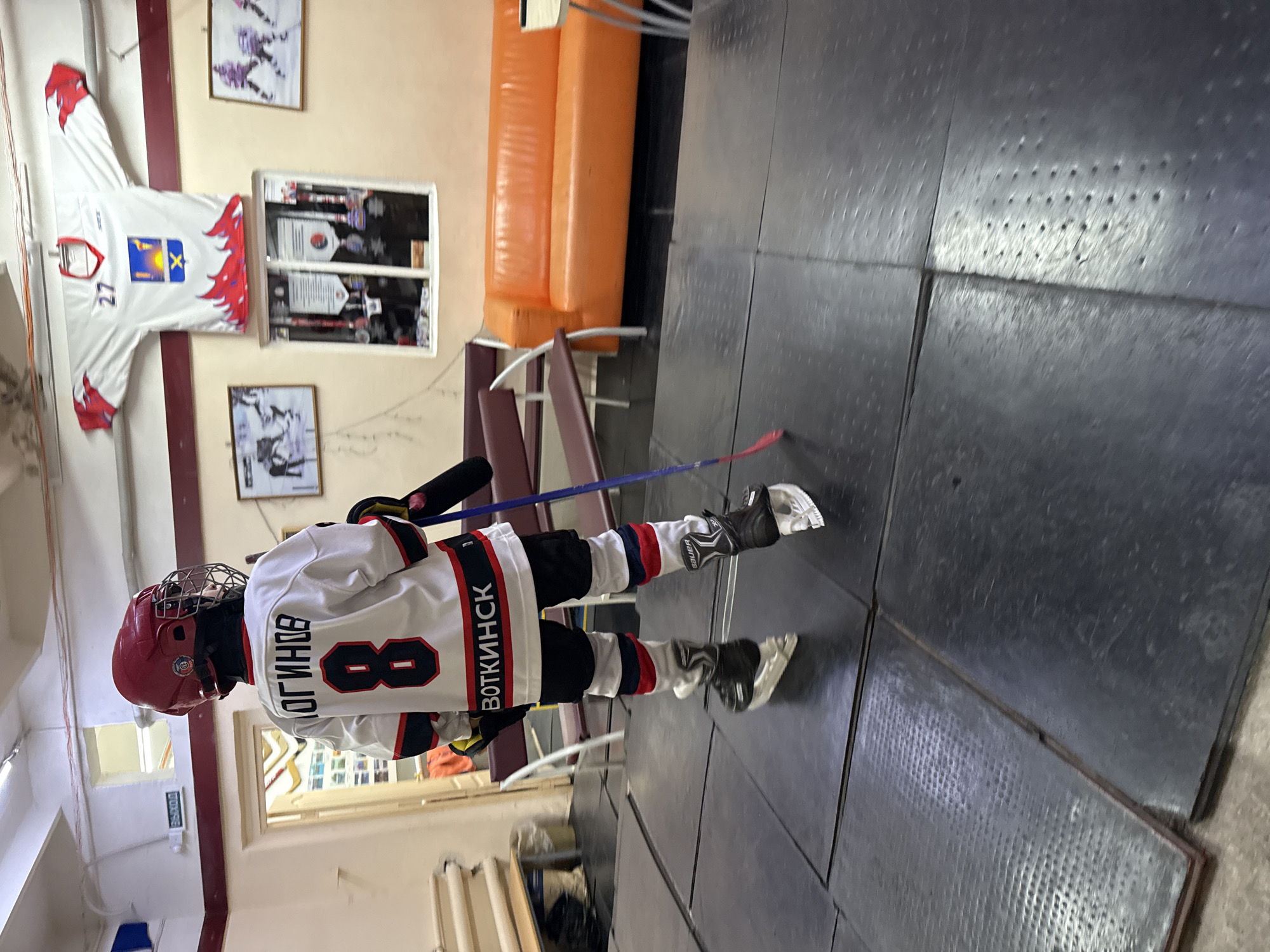 A young person in a hockey uniform, complete with helmet, skates, and protective gear, is standing on a black mat floor. The uniform is white with red and blue accents, featuring the name "ОГИНОВ" and the number "8" on the back, along with "ВОТКИНСК" below. The person is holding a hockey stick.
The background shows a room with a red and blue couch, benches, and framed pictures on the wall, including what appears to be a hockey jersey with the number "27" and a coat of arms. There's also a display case with medals and certificates. A sign with the word "ВЫХОД" (EXIT) is visible above a doorway on the left. The environment suggests a sports facility or locker room, possibly within Votkinsk, Russia, given the text on the uniform. The lighting appears to be artificial, coming from overhead fixtures.