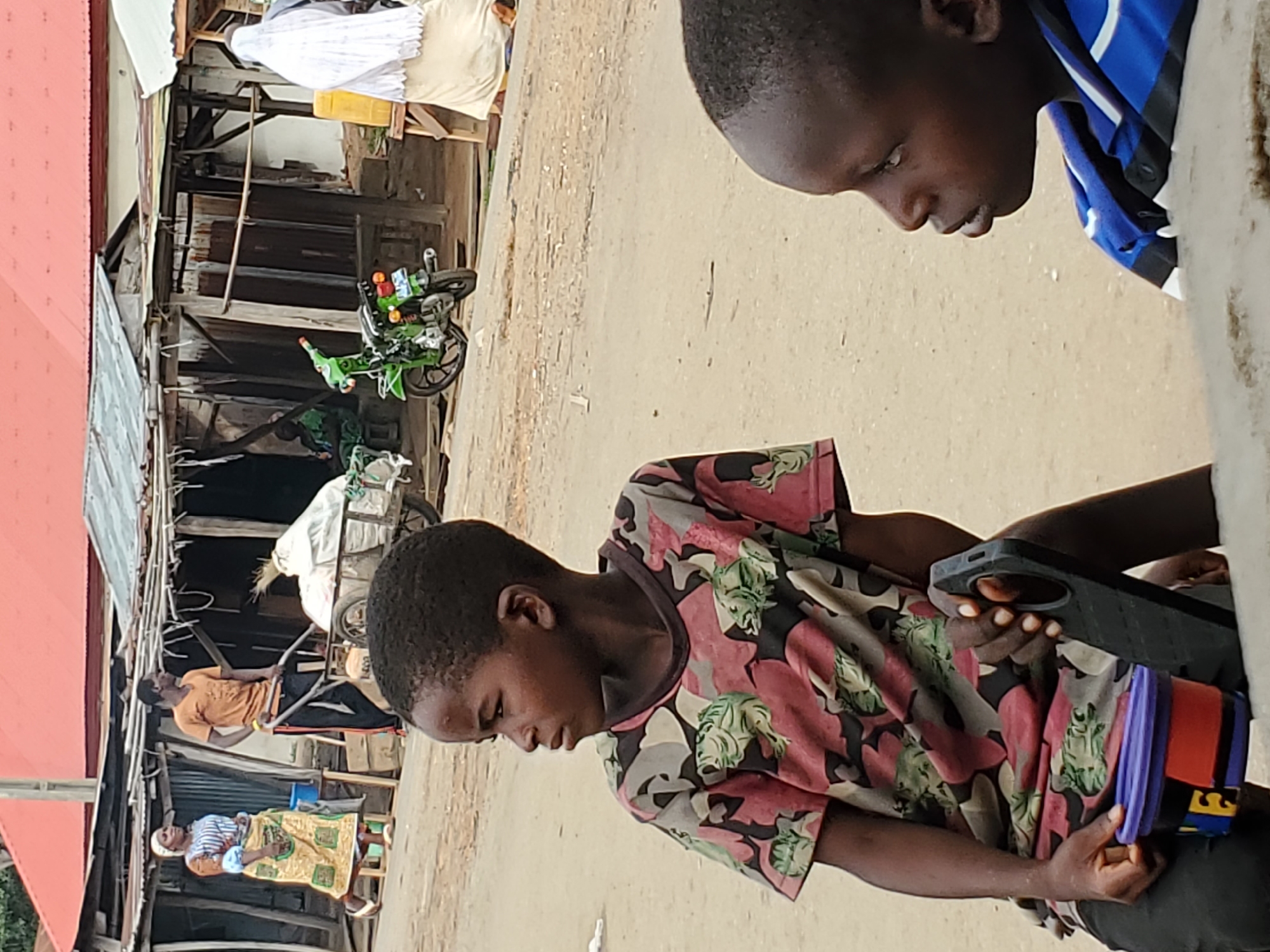 Two young men are in the foreground, one holding a smartphone. Behind them is a dirt road with a building to the left. People are visible near the building, including one pushing a cart. A green motorcycle is parked near the building. The location is Mararaba, Nigeria.