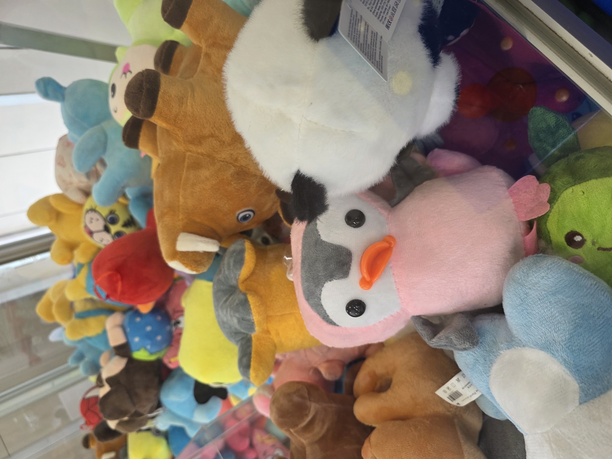 The image depicts a claw machine filled with stuffed animals. The machine is located in Krasnodar, Russia. The toys include various colors and animals, such as a penguin, blue dog, brown horse, red heart, and green alien. The animals are tightly packed together within the machine.